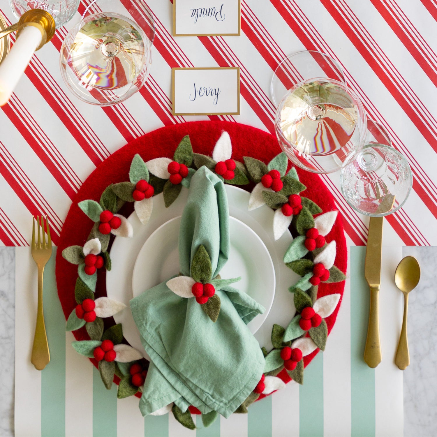 A place setting featuring the Red Felt Placemat, a mint napkin tied in the Felt Berry Napkin Ring and Felt Berry Charger atop, Gold Frame Place Card, and gold flatware on the Candy Stripe Runner and Seafoam Classic Stripe Runner.