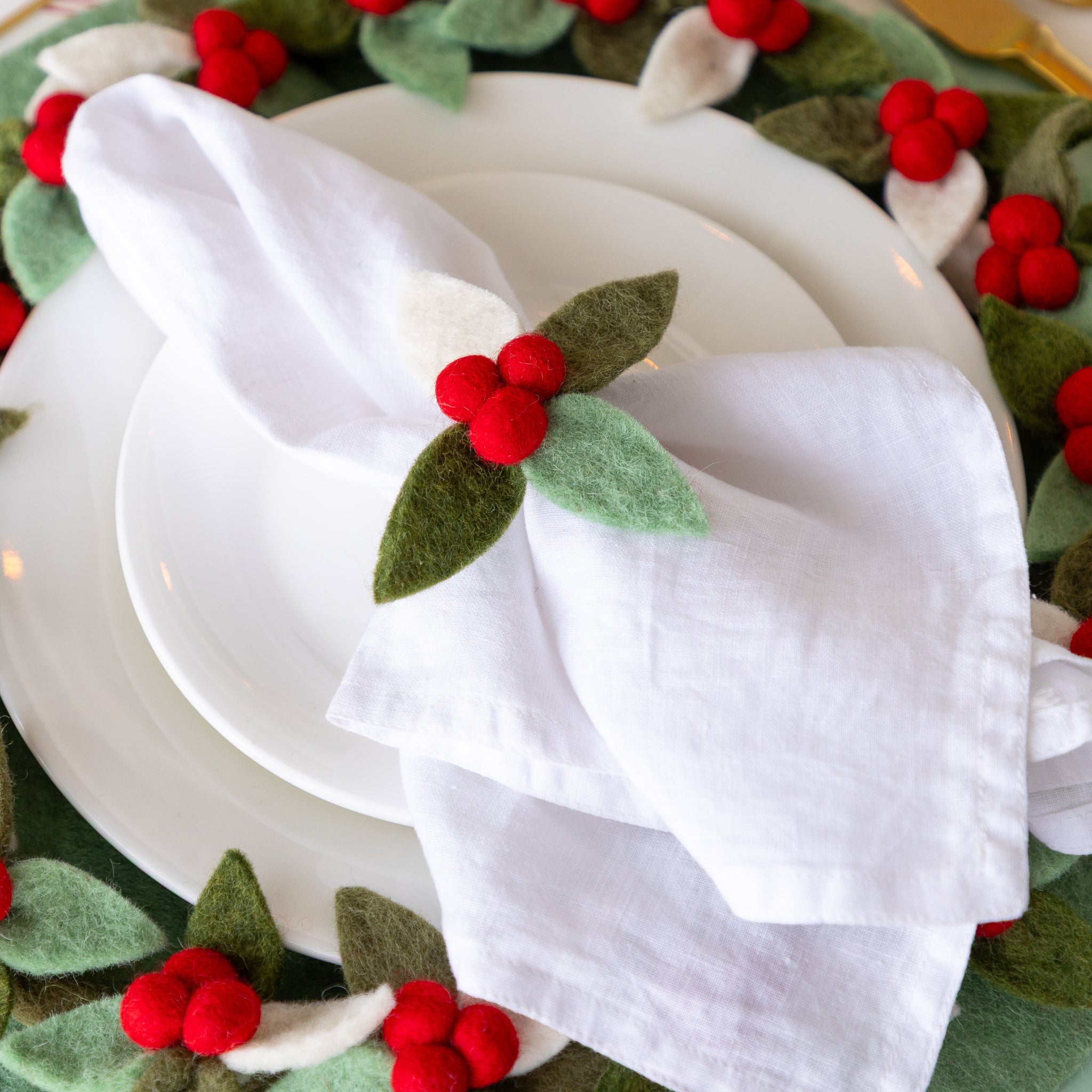 A place setting featuring a white napkin in the Felt Berry Napkin Ring, atop white dinner plates.