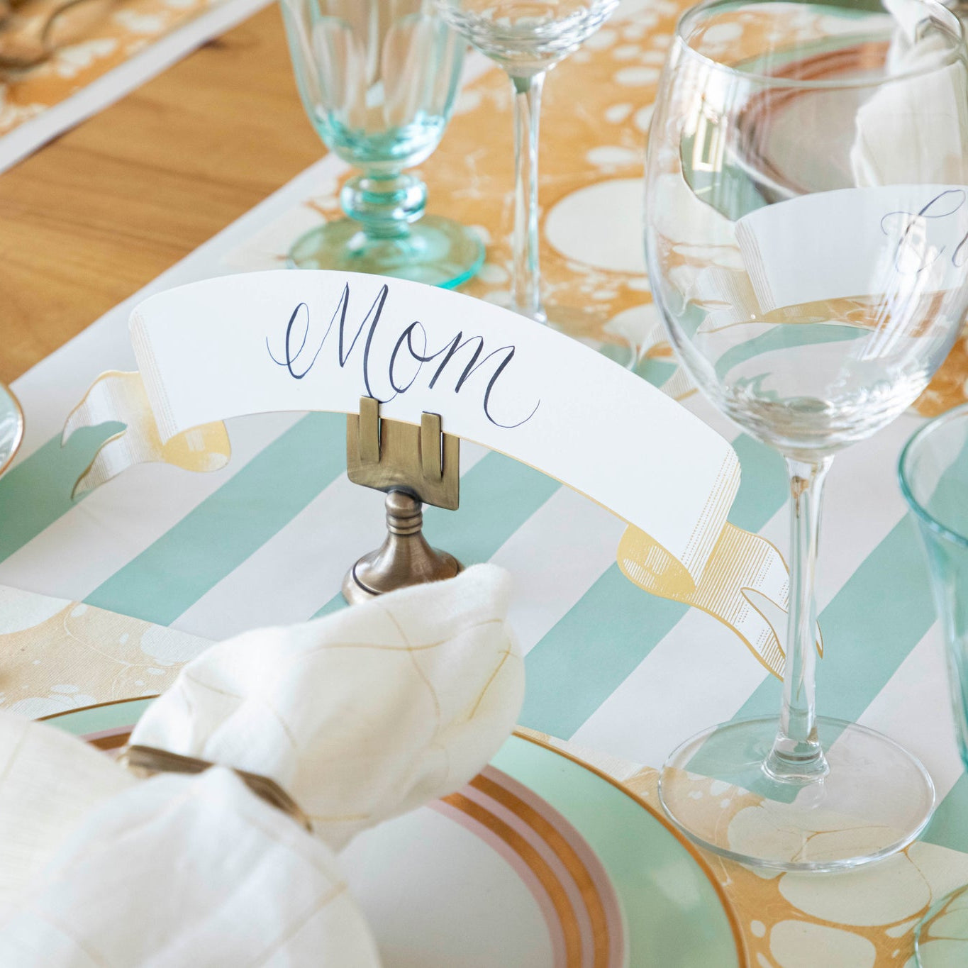 Classic Gold Banner Table Accent with "Mom" written on it, held up by the Brass Place Card holder behind a place setting.
