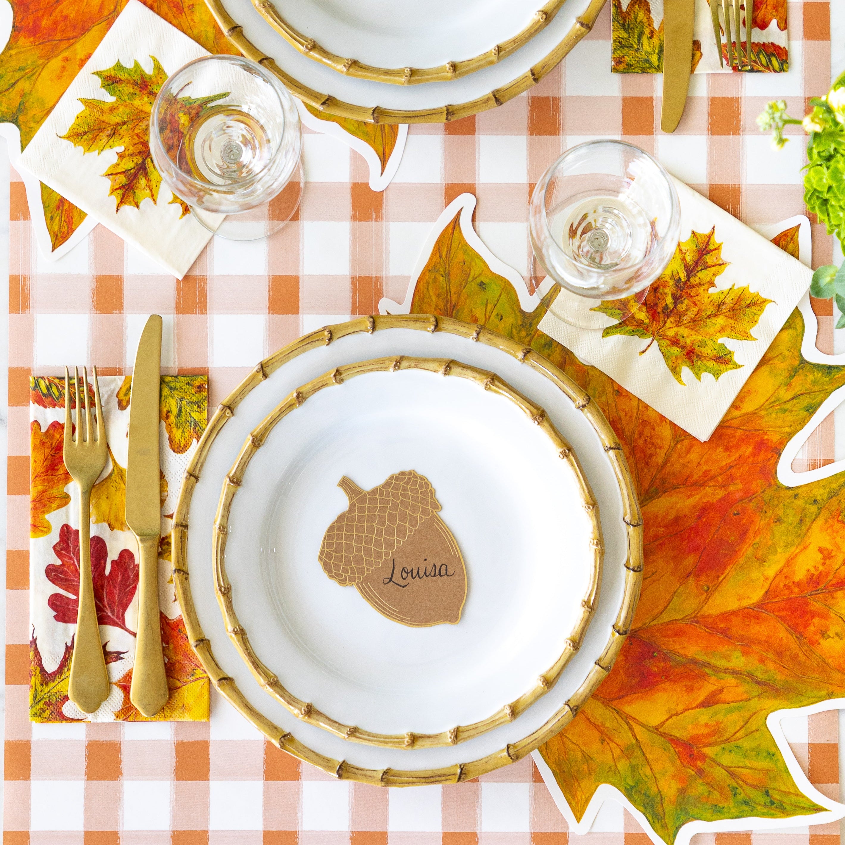 An elegant place setting featuring the Die-cut Fall Leaf Placemat, Juliska Bamboo plates, the Acorn Place Card reading "Louisa", Fall Foliage Cocktail and Guest Napkins and the Orange Painted Check Runner underneath.