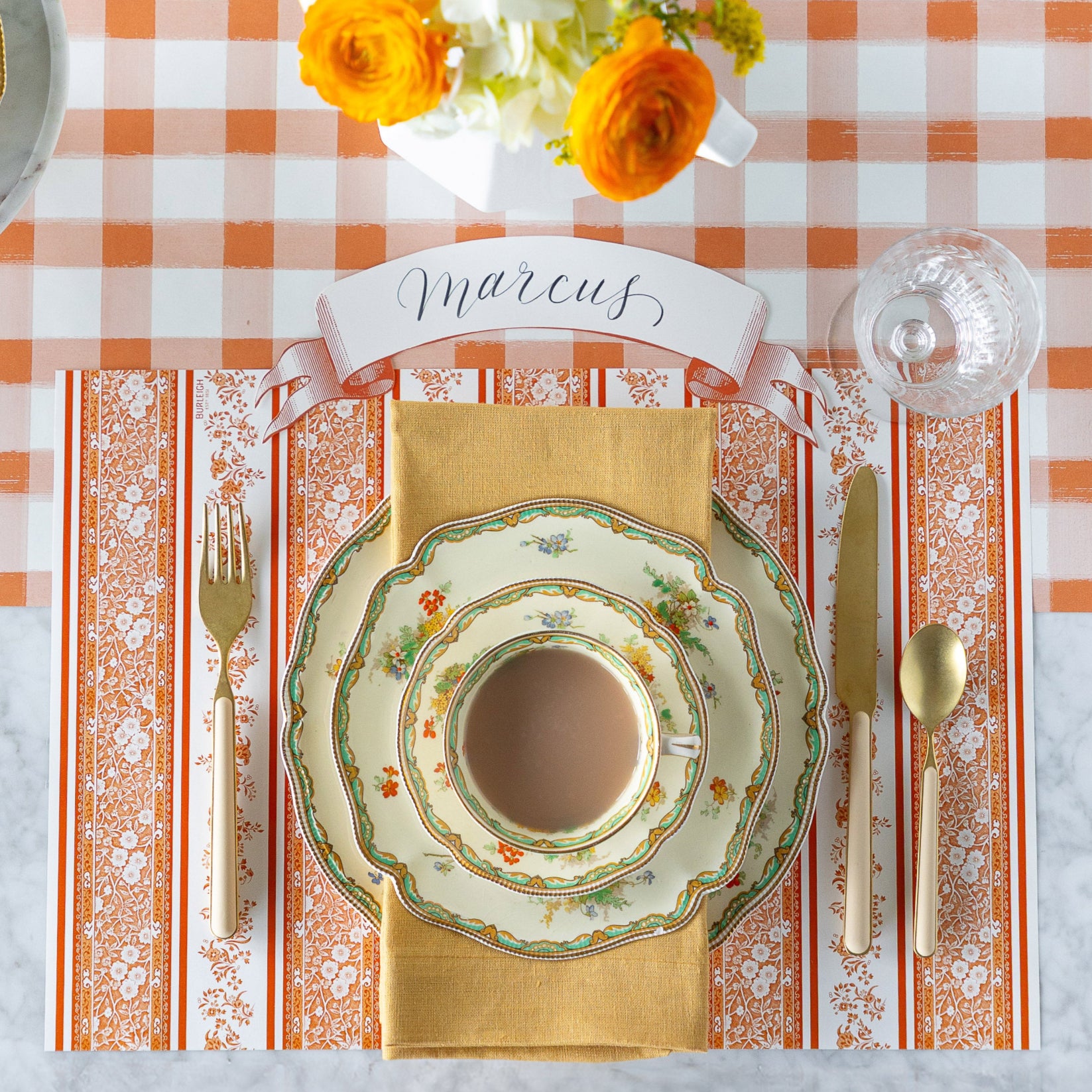 A fall place setting featuring the Pumpkin Burleigh Stripe Placemat, vintage floral dinnerware, a tan napkin, Orange Classic Banner Table Accent reading "Marcus", and gold flatware on the Orange Painted Check Runner.