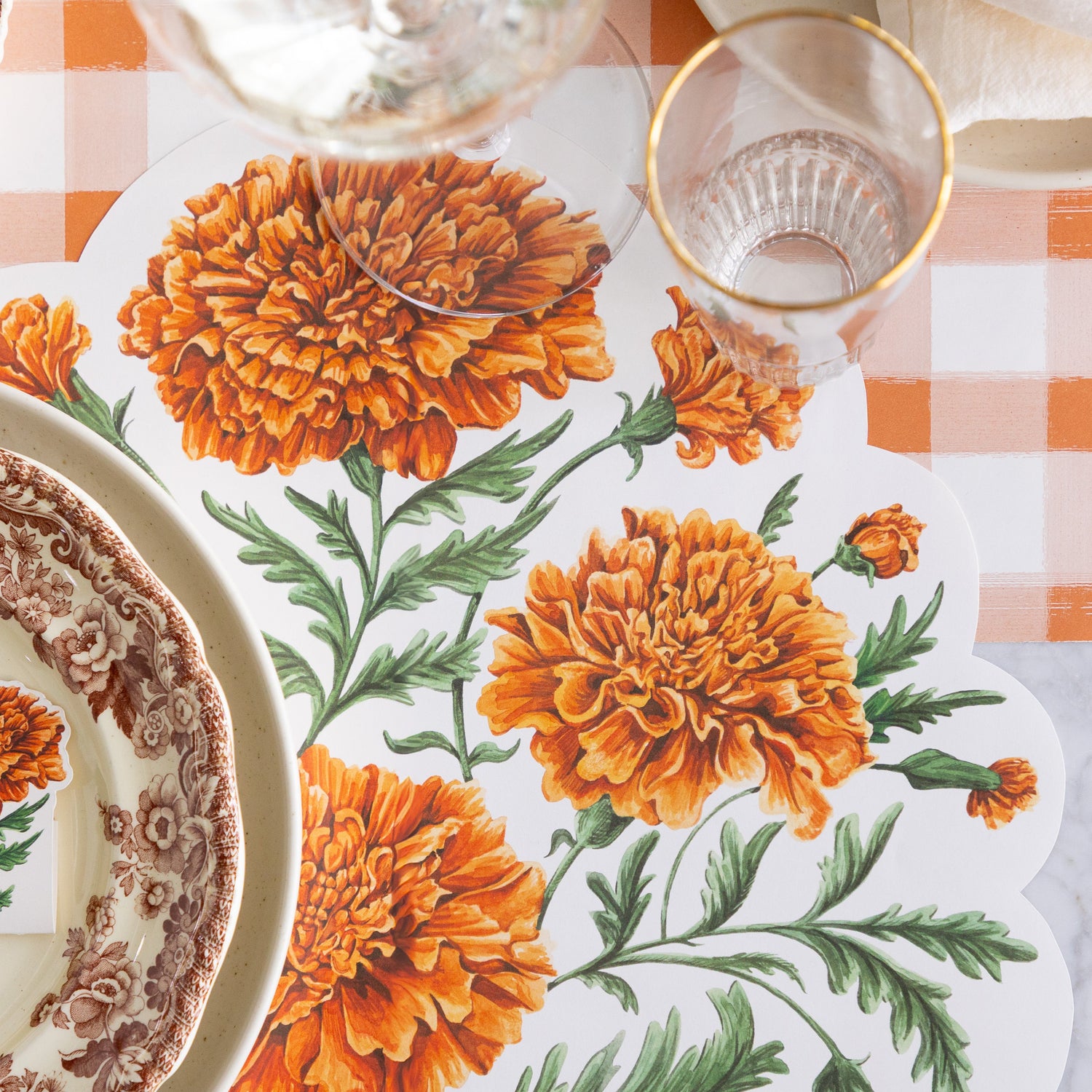 An elegant place setting featuring the Die-cut Marigold Bouquet Placemat offset to the right of the dinner plates, with a wine and water glass on top and Orange Painted Check Runner underneath.