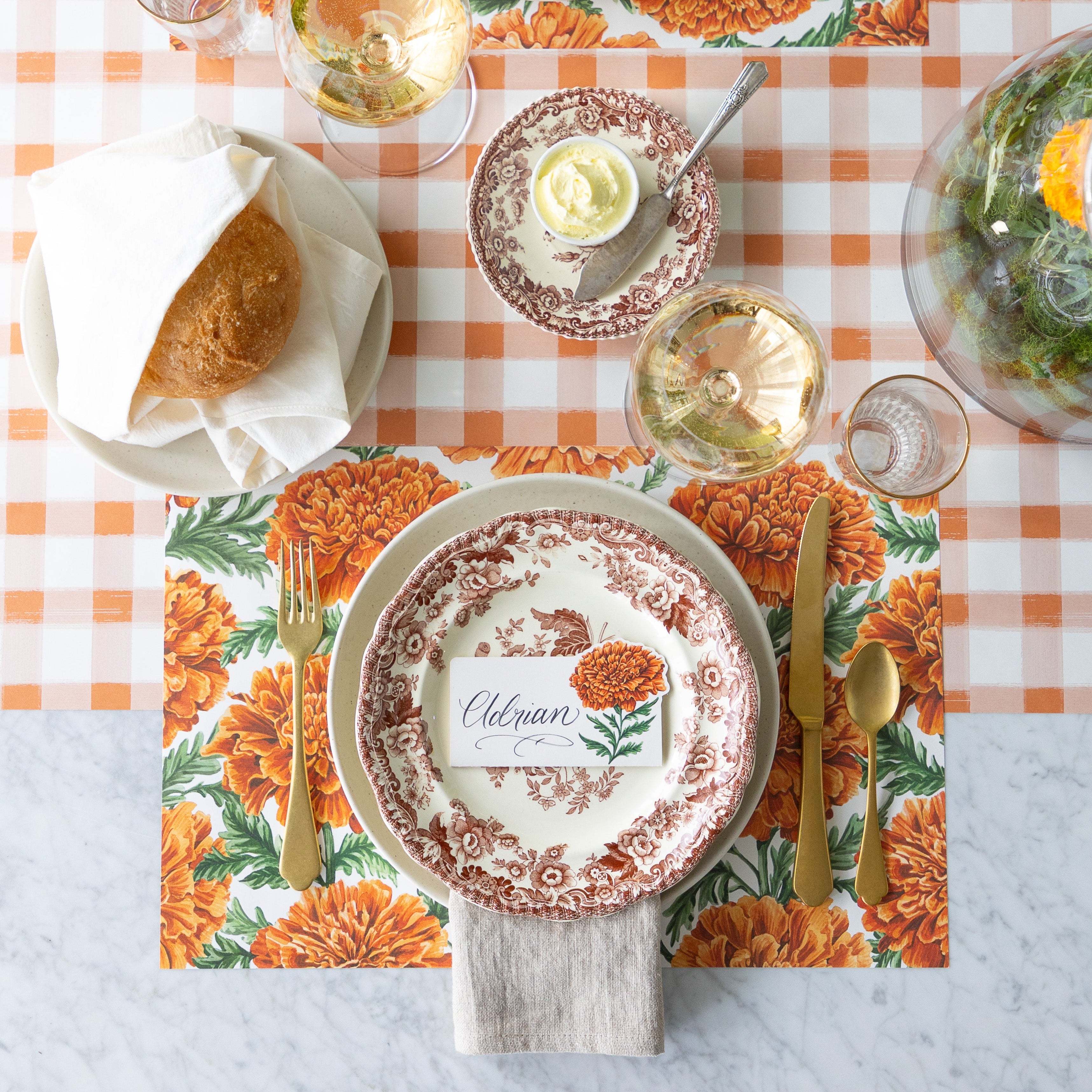 An elegant table setting with a vintage floral plate, gold utensils, bread roll, wine glass, and a Marigold Bloom Place Card reading "Eleanor" atop a Marigold Harvest Placemat—on a Orange Painted Check Runner.