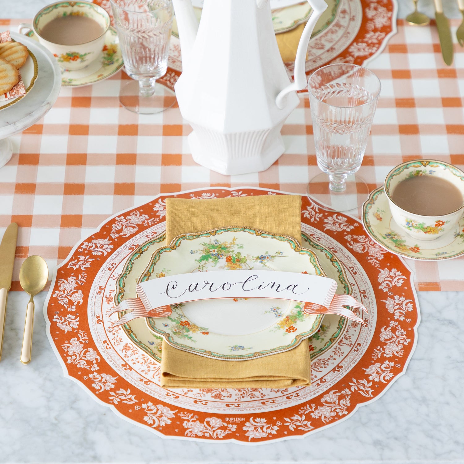 A fall place setting featuring the Die-cut Pumpkin Regal Placemat, vintage floral dinnerware with a tan napkin and Orange Banner Table Accent reading "Carolina", atop, and a cup of coffee on the Orange Painted Check Runner.