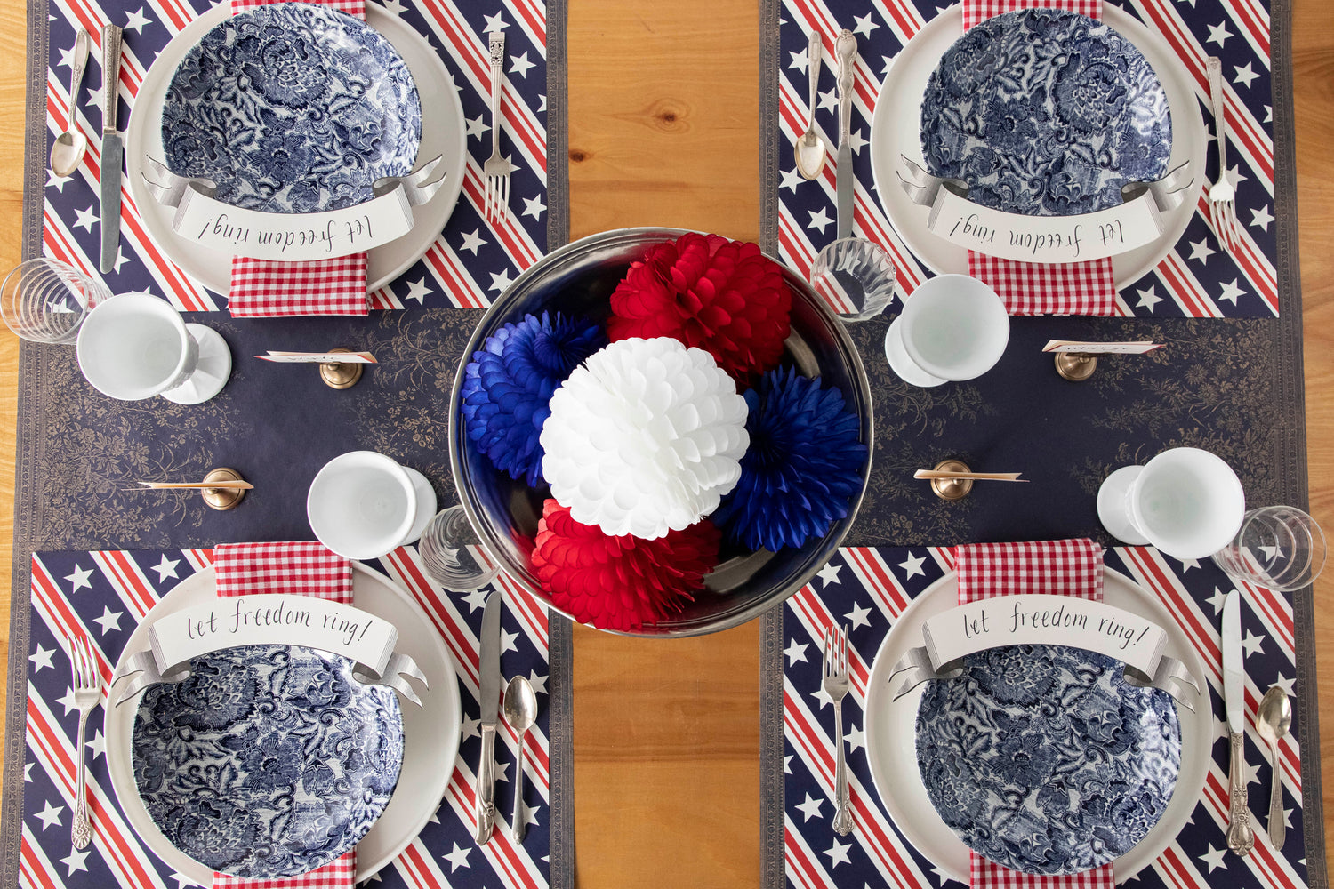 A patriotic themed table setting featuring the Stars & Stripes Placemat with white and blue floral dinner plates, red-and-white checkered napkins, Black Classic Banner Table Accents, and vintage silverware on the Navy Woven Floral Runner.