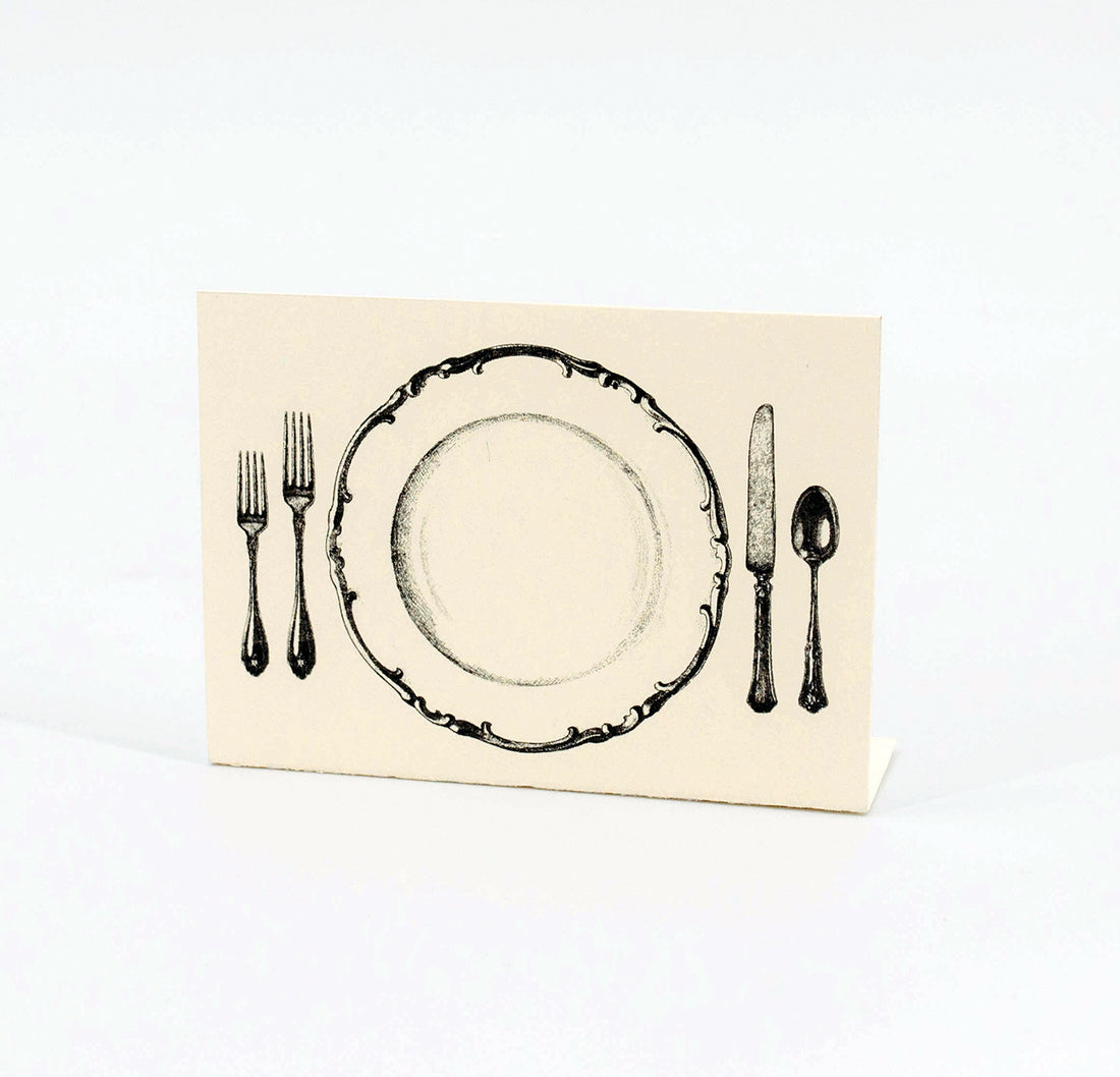 A white, rectangular freestanding place card featuring black artwork of an ornate plate with cutlery on either side.