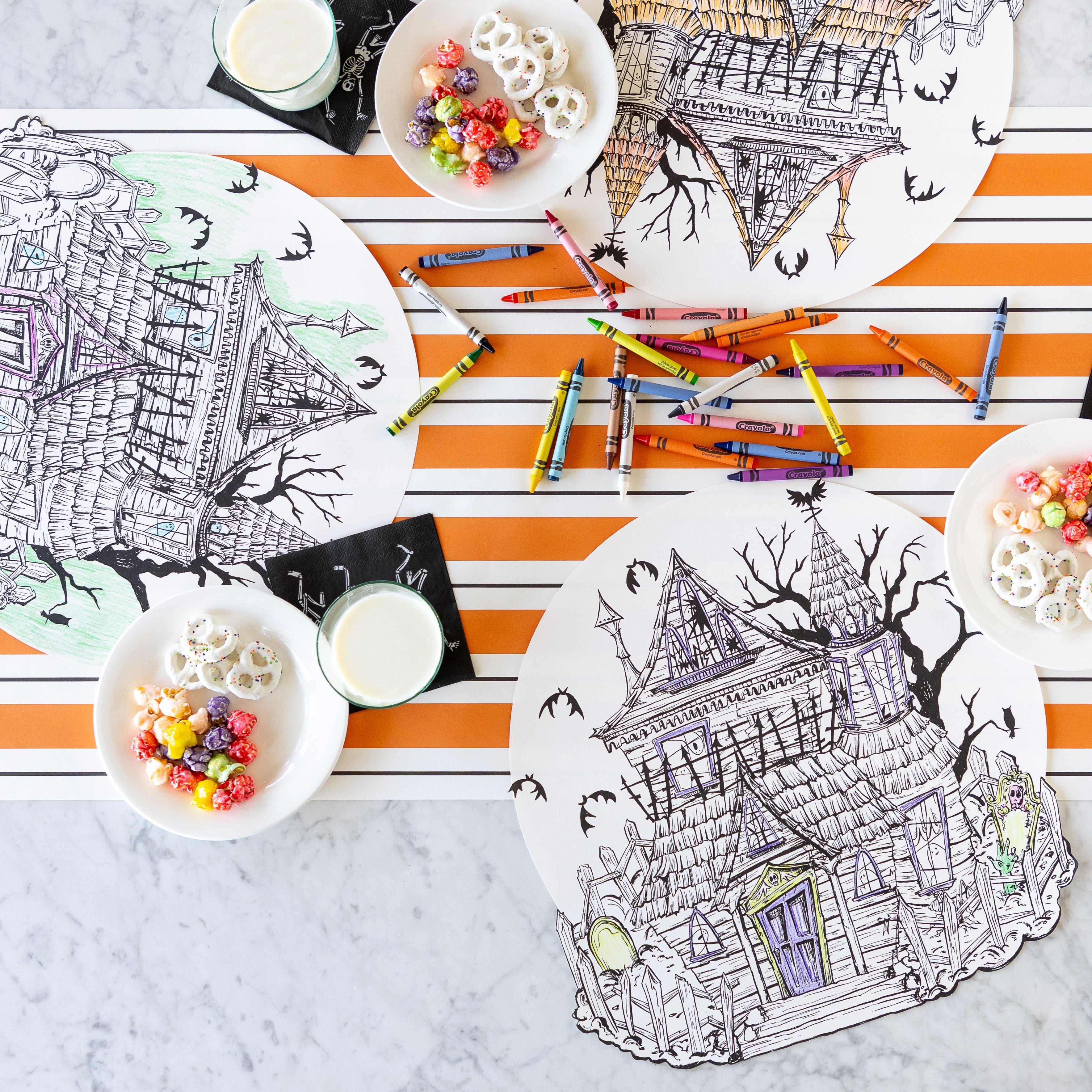 Three Die-cut Haunted House Coloring Placemats partially colored in, surrounded by crayons, glasses of milk on the Skeletons Cocktail Napkins and a bowl of treats with the Orange & Black Awning Stripe Runner underneath.