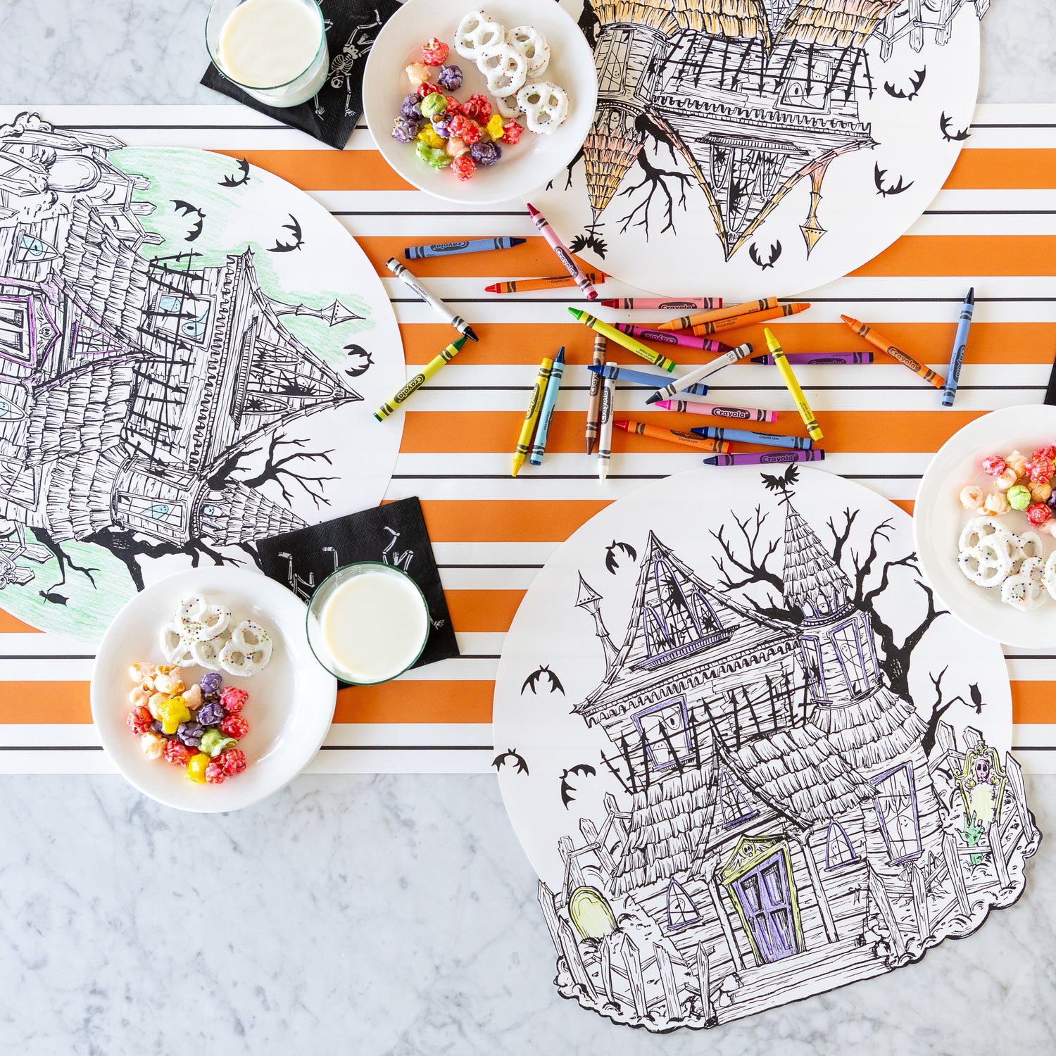 Three Die-cut Haunted House Coloring Placemats partially colored in, surrounded by crayons, glasses of milk on the Skeletons Cocktail Napkins and a bowl of treats with the Orange &amp; Black Awning Stripe Runner underneath.