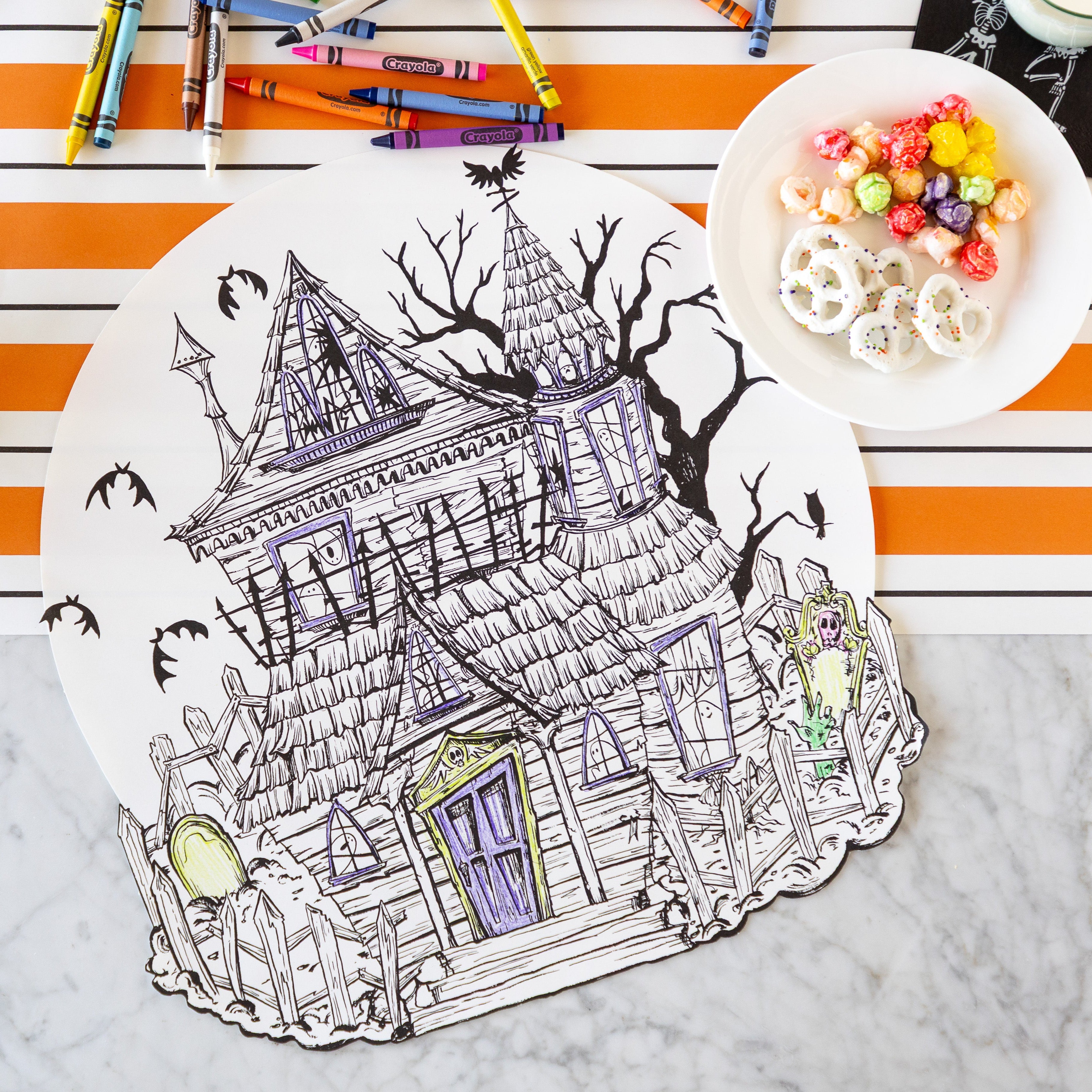 The Die-cut Haunted House Coloring Placemat partially colored in, surrounded by crayons and a bowl of treats, with the Orange & Black Awning Stripe Runner underneath.