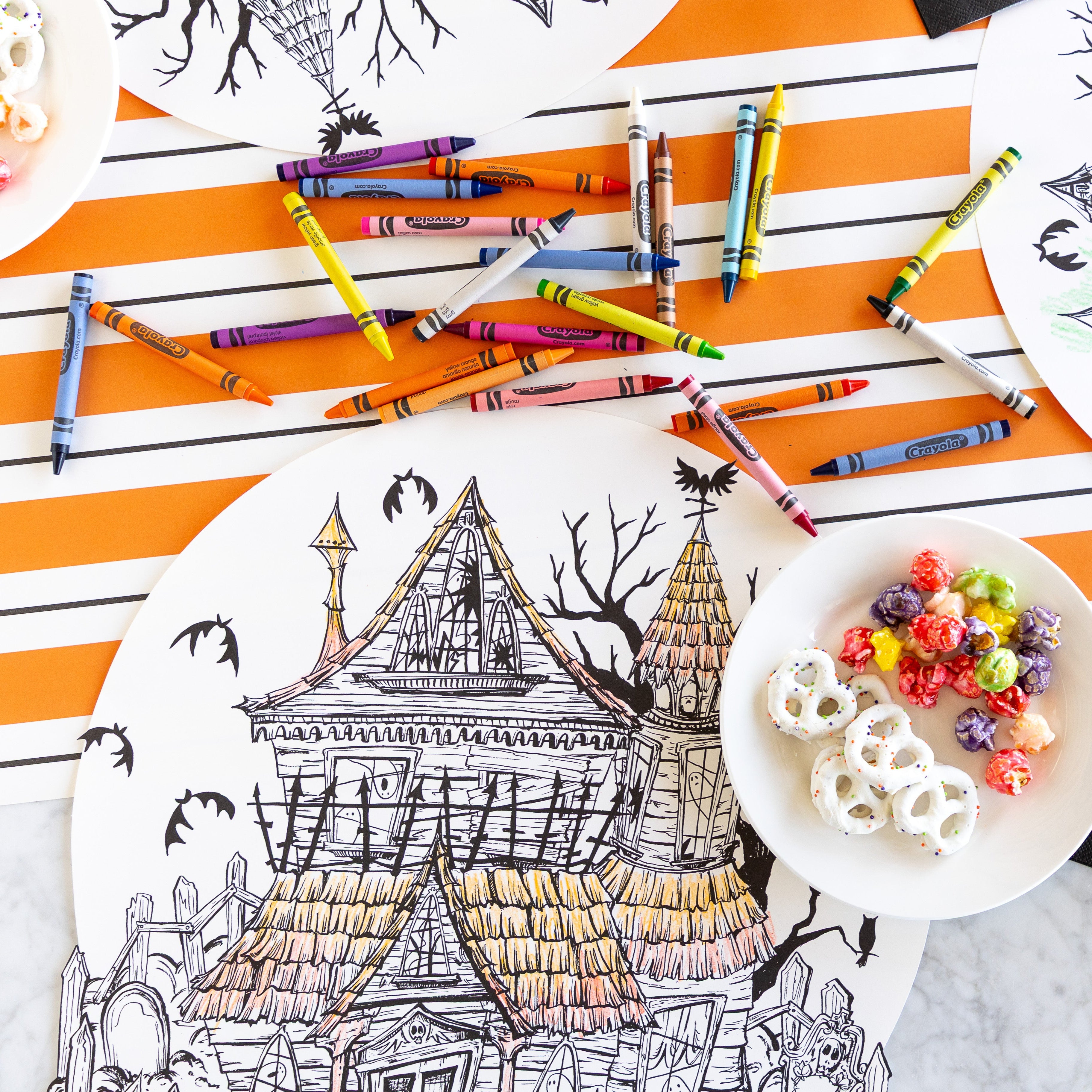 The Die-cut Haunted House Coloring Placemat partially colored in, surrounded by crayons and a bowl of treats, with the Orange & Black Awning Stripe Runner underneath.