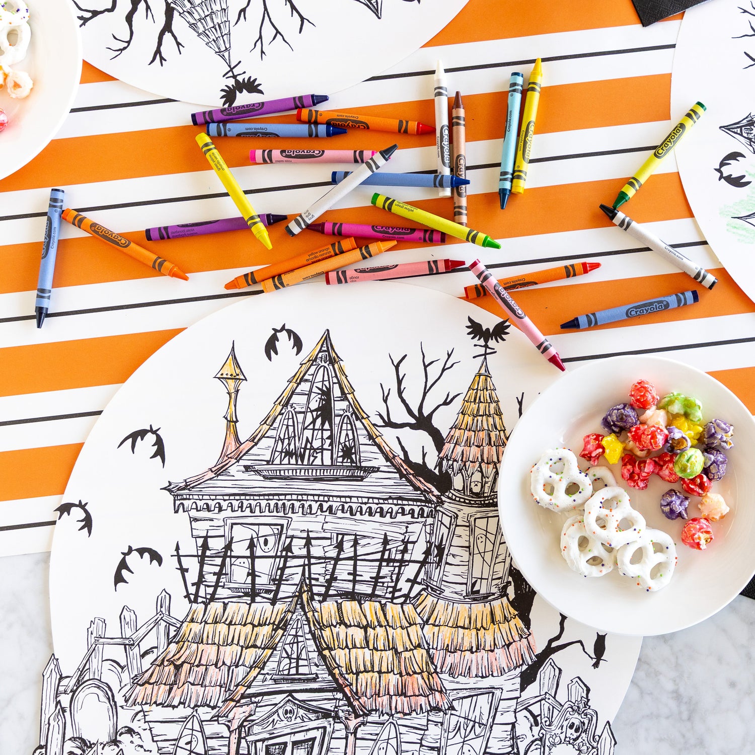 The Die-cut Haunted House Coloring Placemat partially colored in, surrounded by crayons and a bowl of treats, with the Orange &amp; Black Awning Stripe Runner underneath.