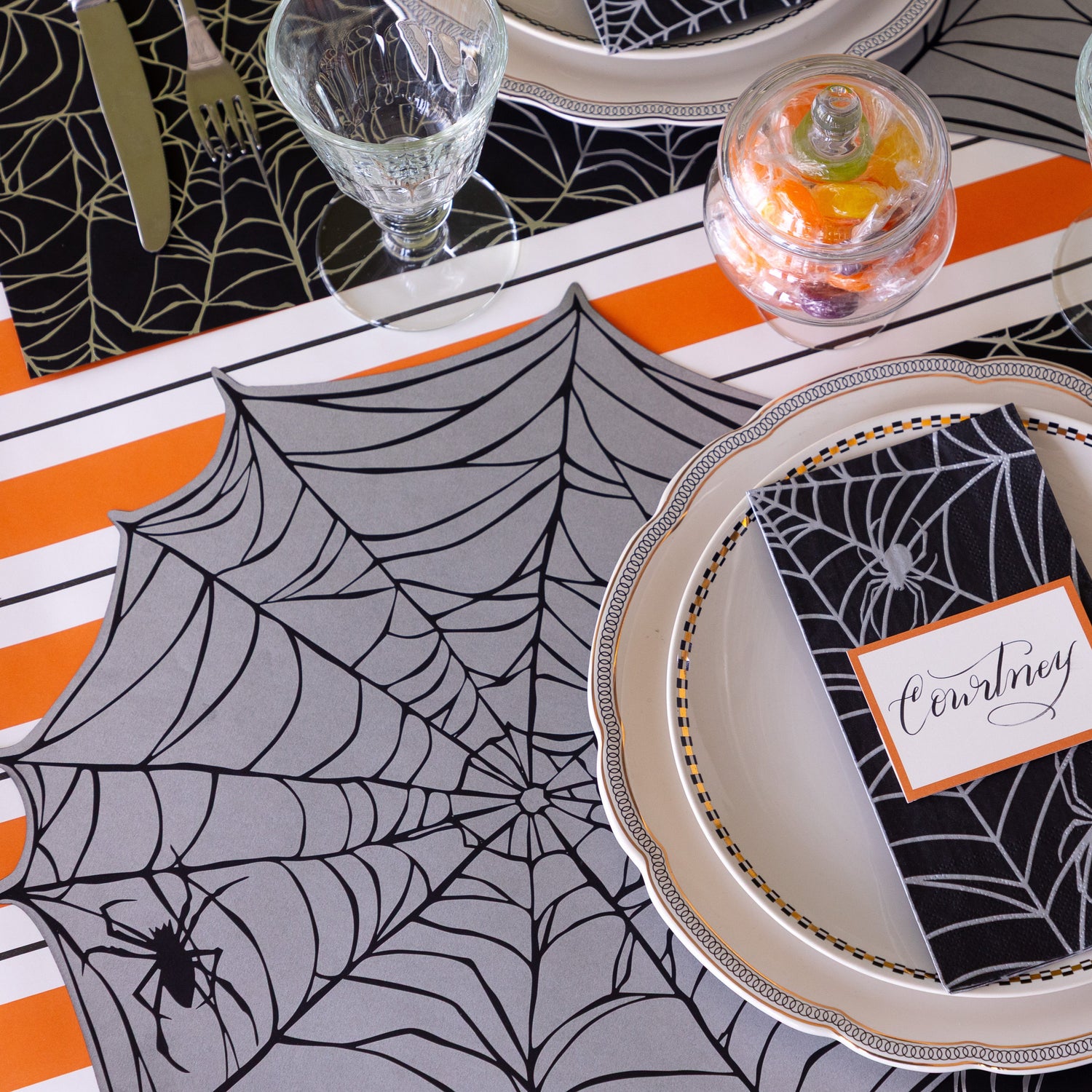 A Halloween themed table setting featuring the Die-cut Spiderweb Placemat,  Spiderweb Guest Napkin on the plates with the Orange Frame Place Card on top, and Orange &amp; Black Awning Stripe Runner underneath.