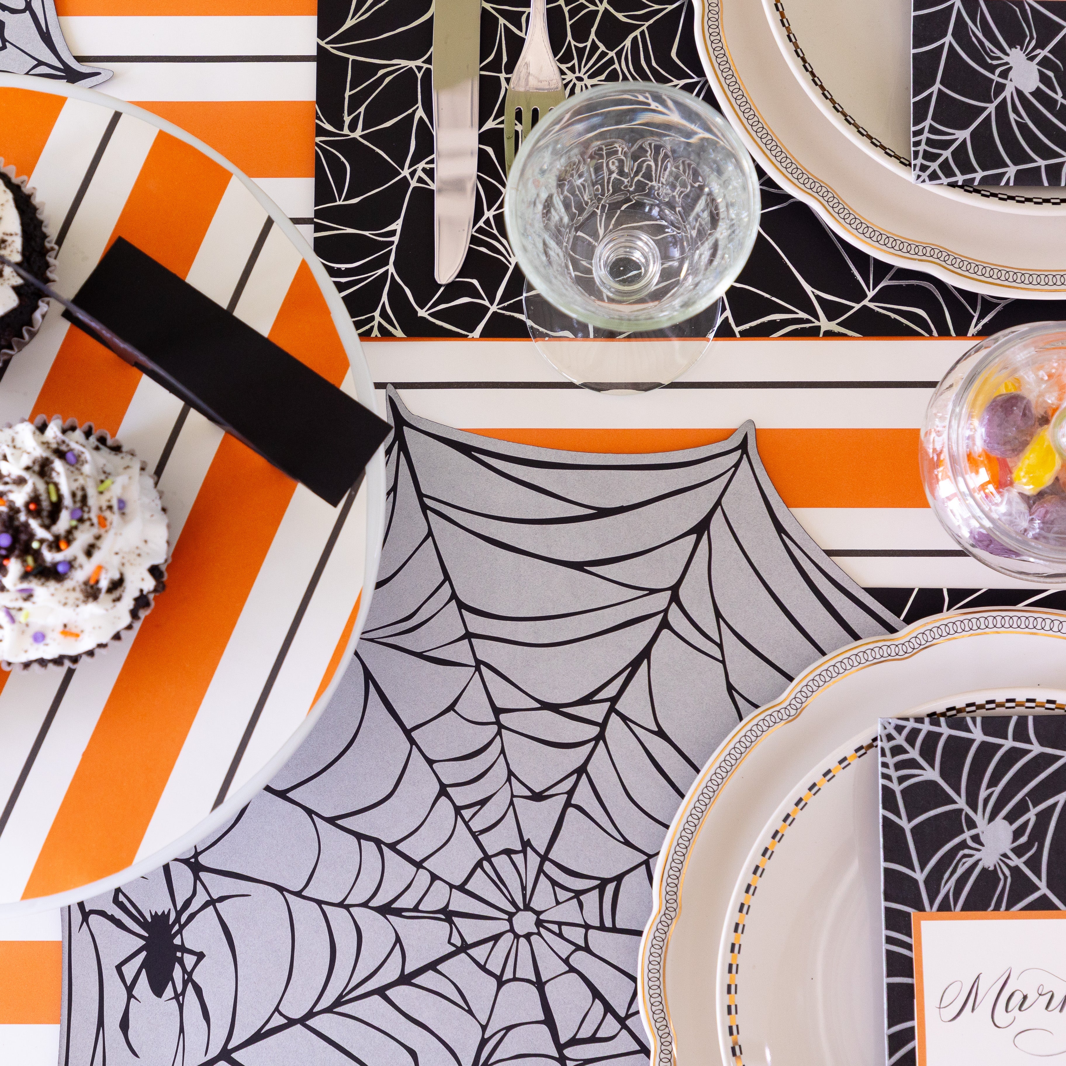 A Halloween themed table setting with halloween themed cupcakes and candy, featuring the Die-cut Spiderweb Placemat on the Orange & Black Awning Stripe Runner.