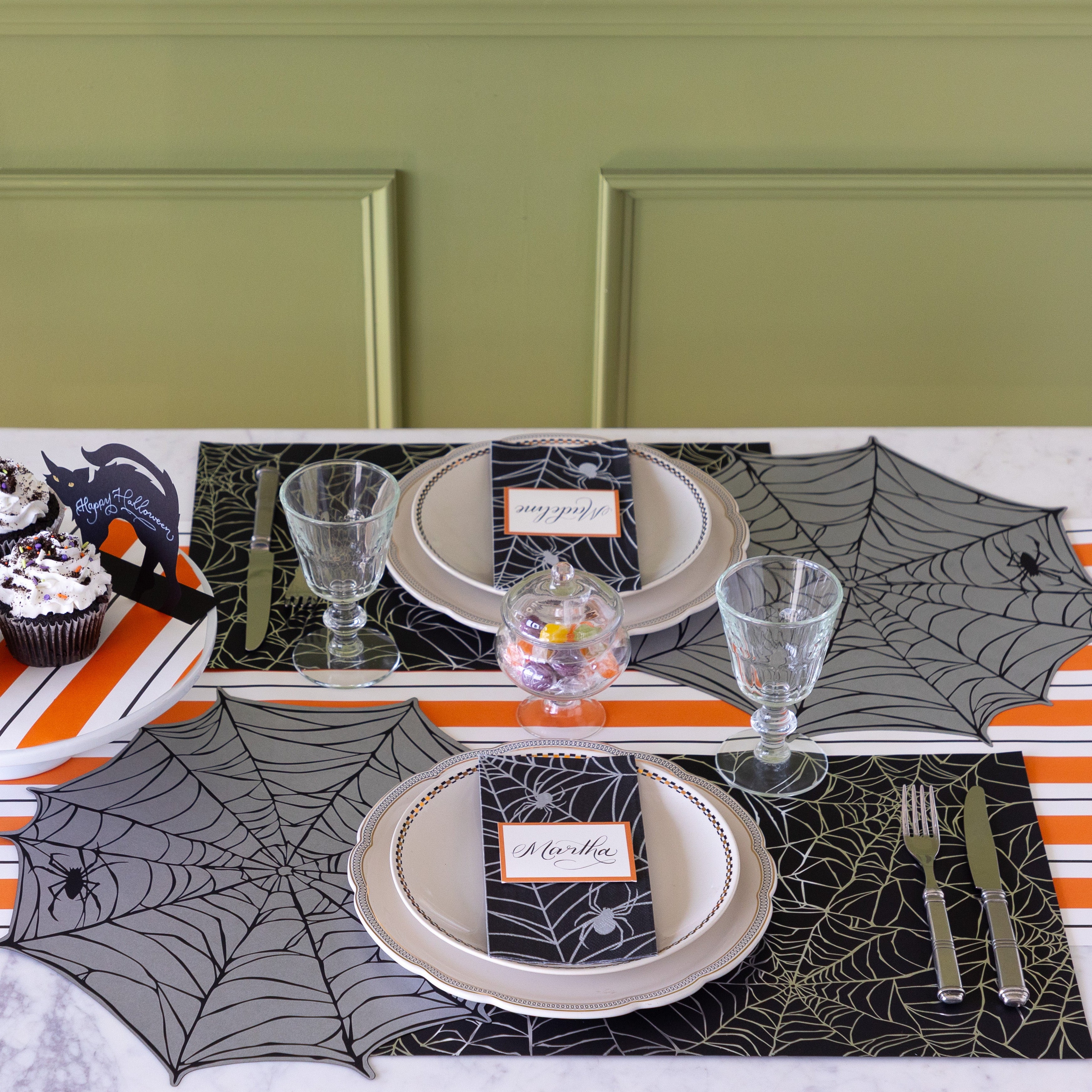 A Halloween themed table setting featuring the Die-cut Spiderweb Placemat slightly overlapping the Spiderweb Placemat, the Spiderweb Guest Napkin with Orange Frame Place Card on top and Orange & Black Awning Stripe Runner underneath.