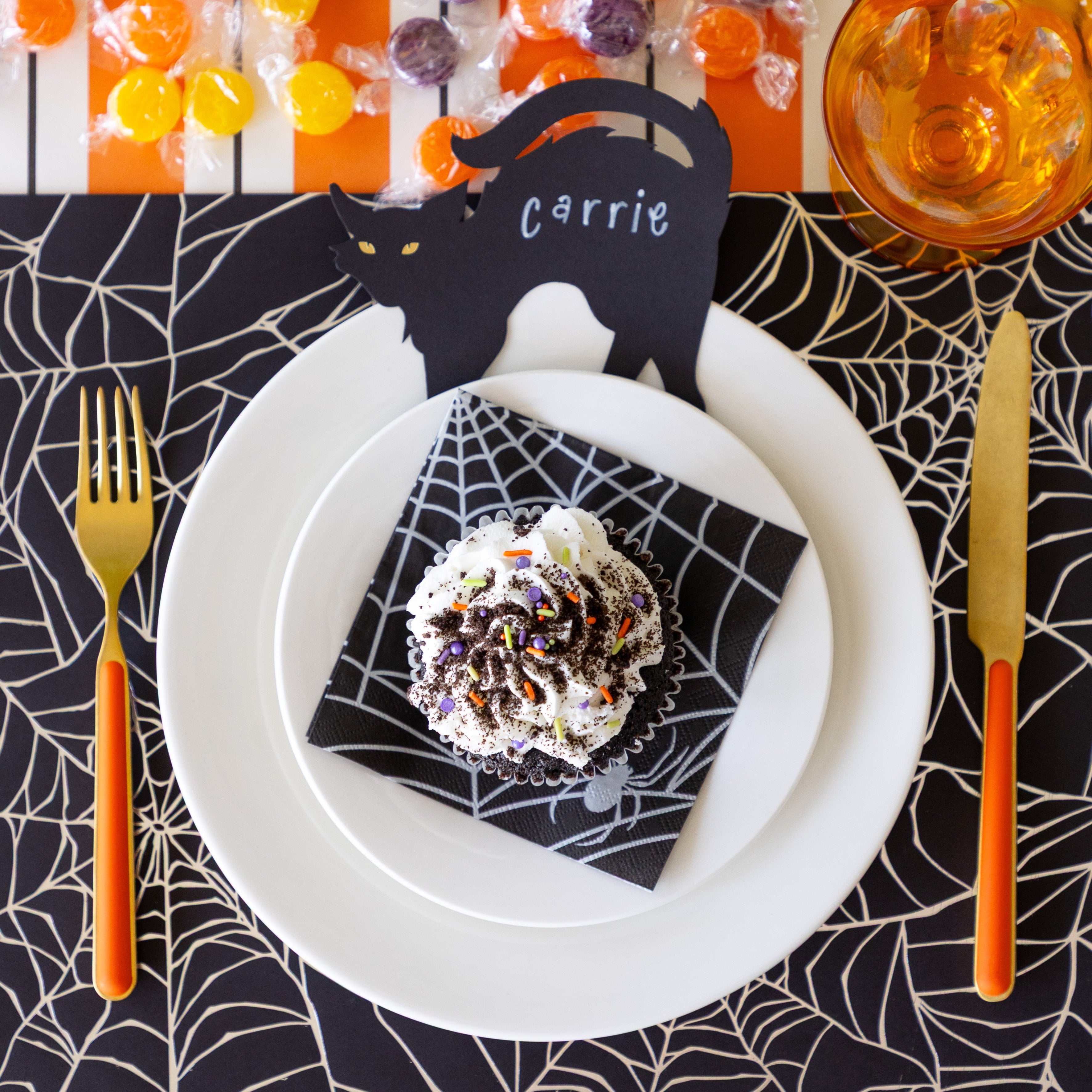 A Halloween place setting featuring the Spiderweb Placemat with a cupcake on the Spiderweb Cocktail Napkin on the plates, and Black Cat Place Card reading "Carrie".