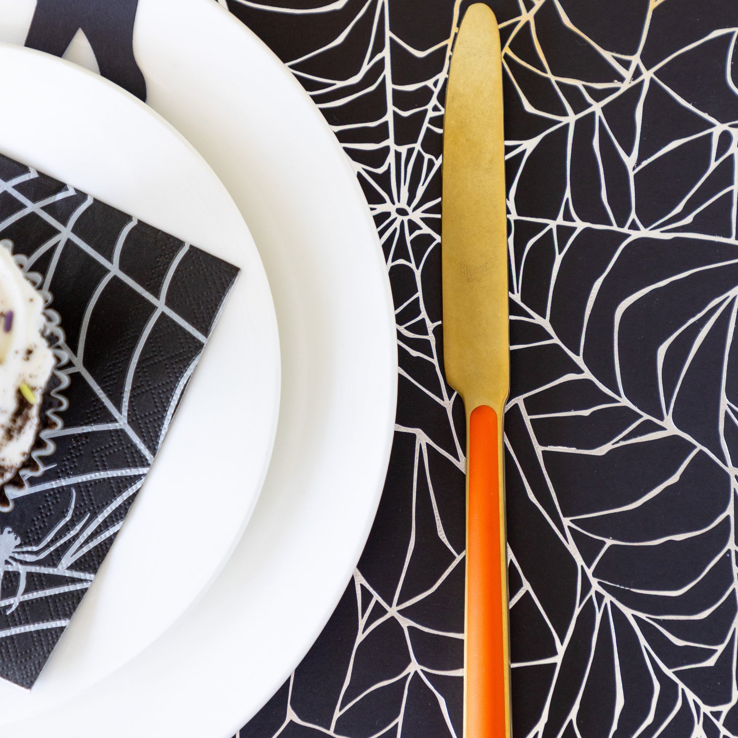 A place setting featuring the Spiderweb Placemat with an orange stemmed, gold knife, white dinner plates and Spiderweb Cocktail Napkin on top.
