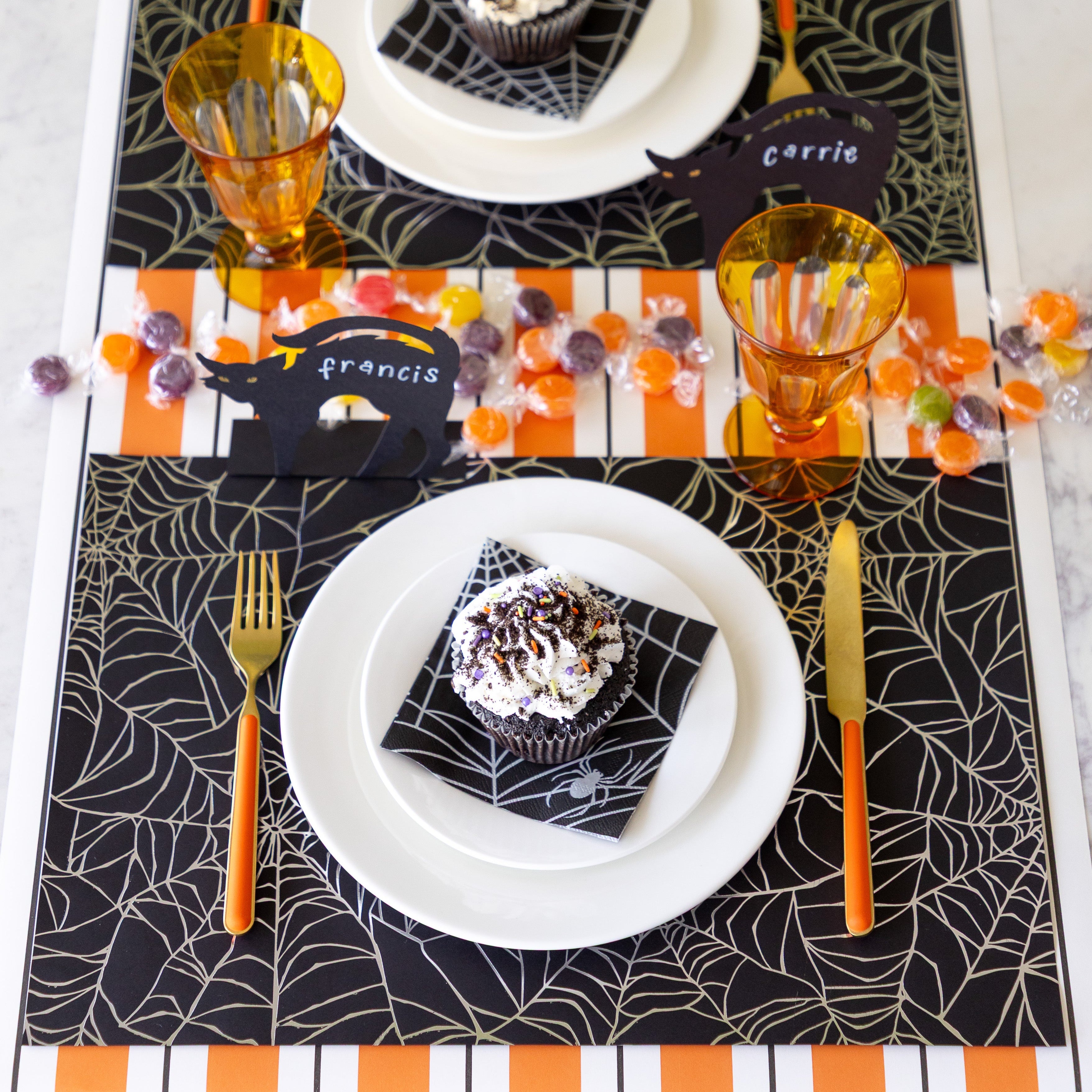A Halloween table setting with candy featuring the Spiderweb Placemat with an orange stemmed, gold knife and fork, a cupcake on the Spiderweb Cocktail Napkin resting on the plates, Black Cat Place Card reading "Francis" and Orange & Black Awning Stripe Runner underneath.