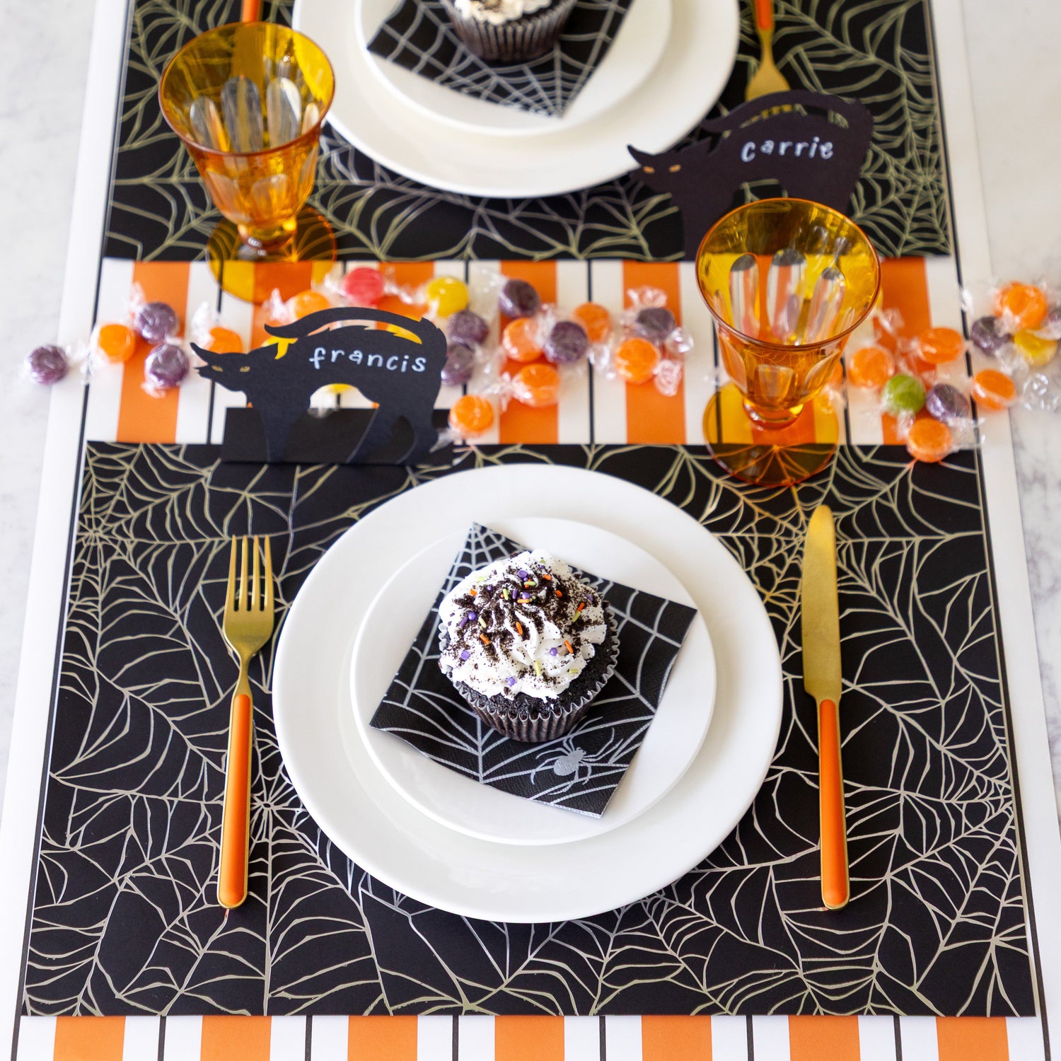 A Halloween table setting with candy featuring the Spiderweb Placemat with an orange stemmed, gold knife and fork, a cupcake on the Spiderweb Cocktail Napkin resting on the plates, Black Cat Place Card reading &quot;Francis&quot; and Orange &amp; Black Awning Stripe Runner underneath.