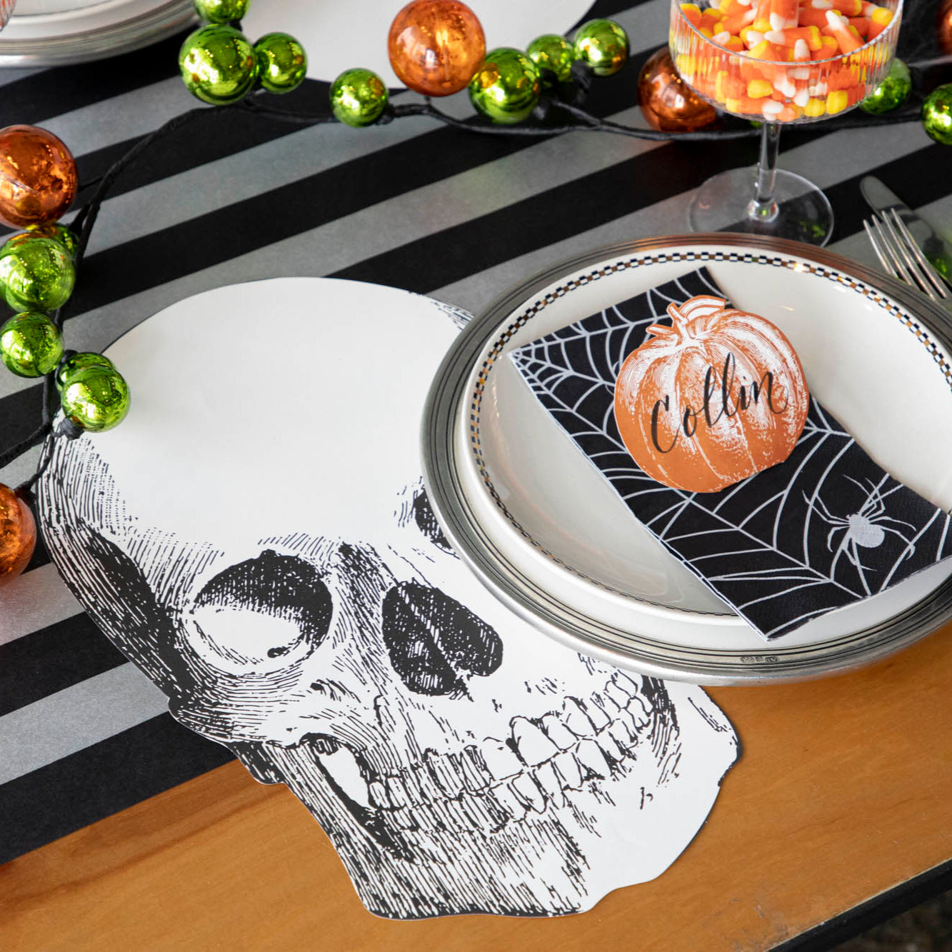 A spooky table setting featuring the Die-cut Skull Placemat, offset to the left of a place setting with the Spiderweb Guest Napkin and Pumpkin Place Card atop.