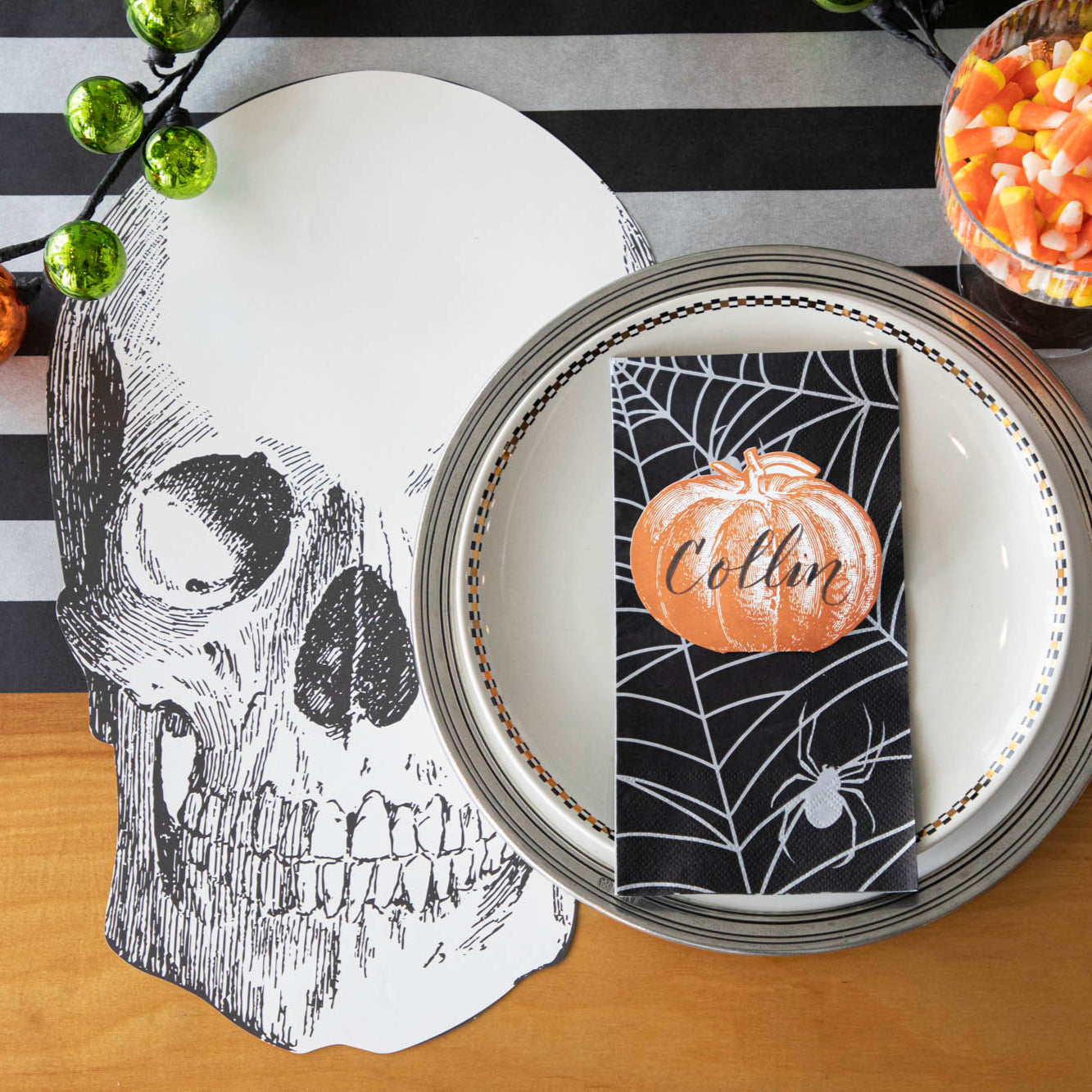A spooky table setting featuring the Die-cut Skull Placemat, offset to the left of a place setting with the Spiderweb Guest Napkin and Pumpkin Place Card atop.