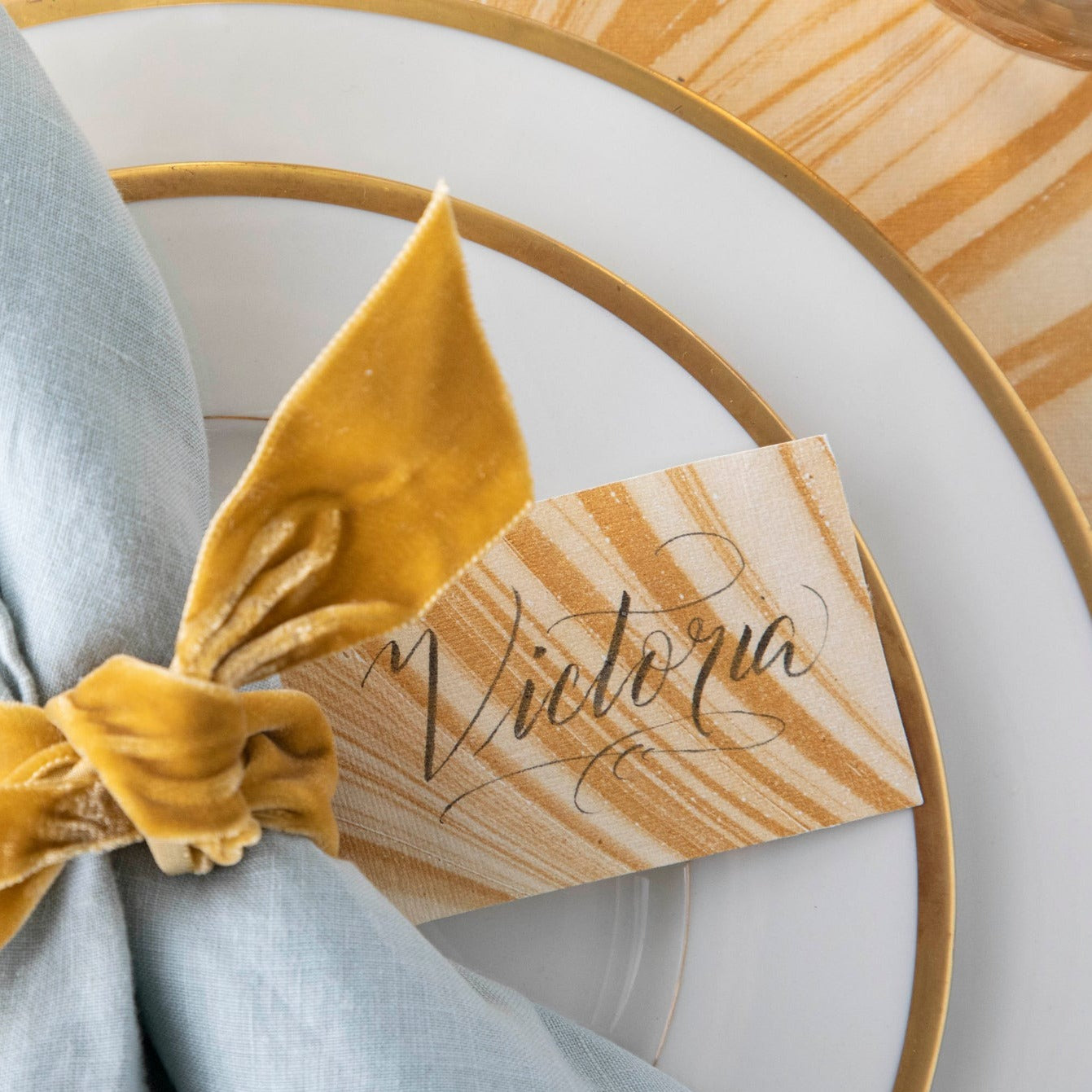 A Gold and cream gift tag tied to the napkin at a place setting, labeled &quot;Victoria&quot;.