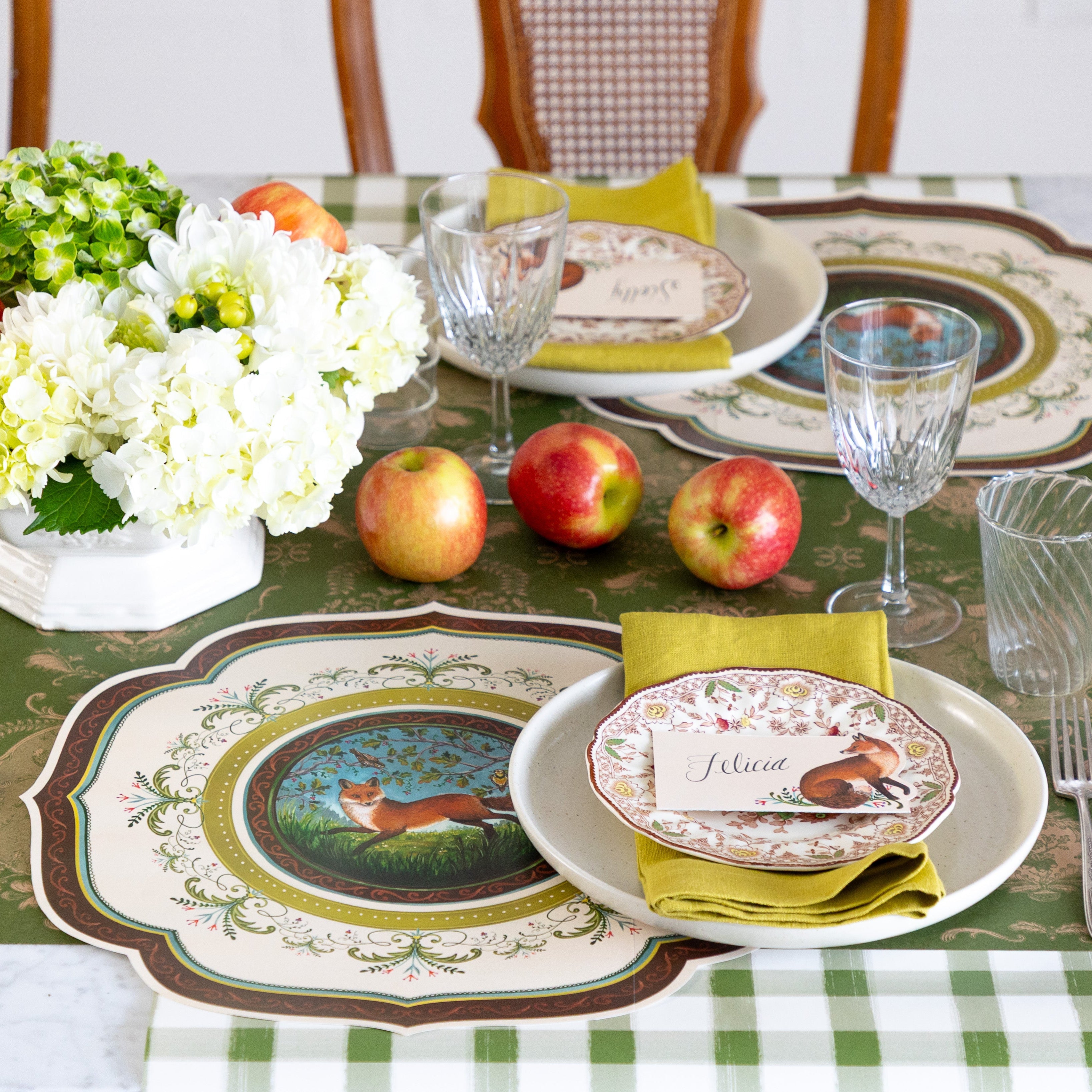 A decorative table setting with scattered apples and flowers, featuring the Die-cut Fanciful Fox China Placemat, vintage dinnerware with the Fanciful Fox Place Card on top and Moss Fable Toile Runner running horizontally and Dark Green Painted Check Runner running vertically on the table.