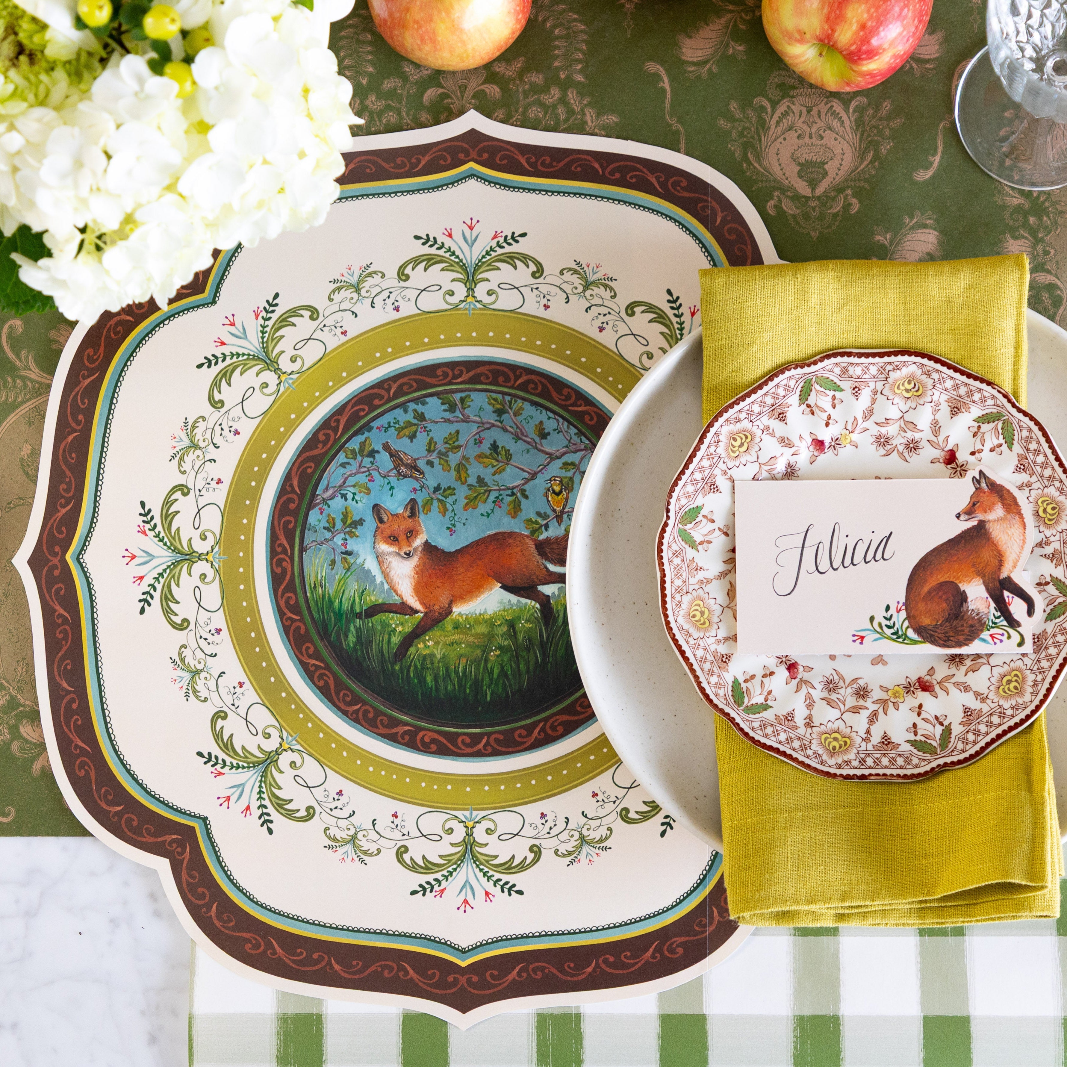 A decorative table setting featuring the Die-cut Fanciful Fox China Placemat, vintage dinnerware with the Fanciful Fox Place Card reading "Felicia" on top and Moss Fable Toile Runner running horizontally and Moss Painted Check Runner running vertically on the table.