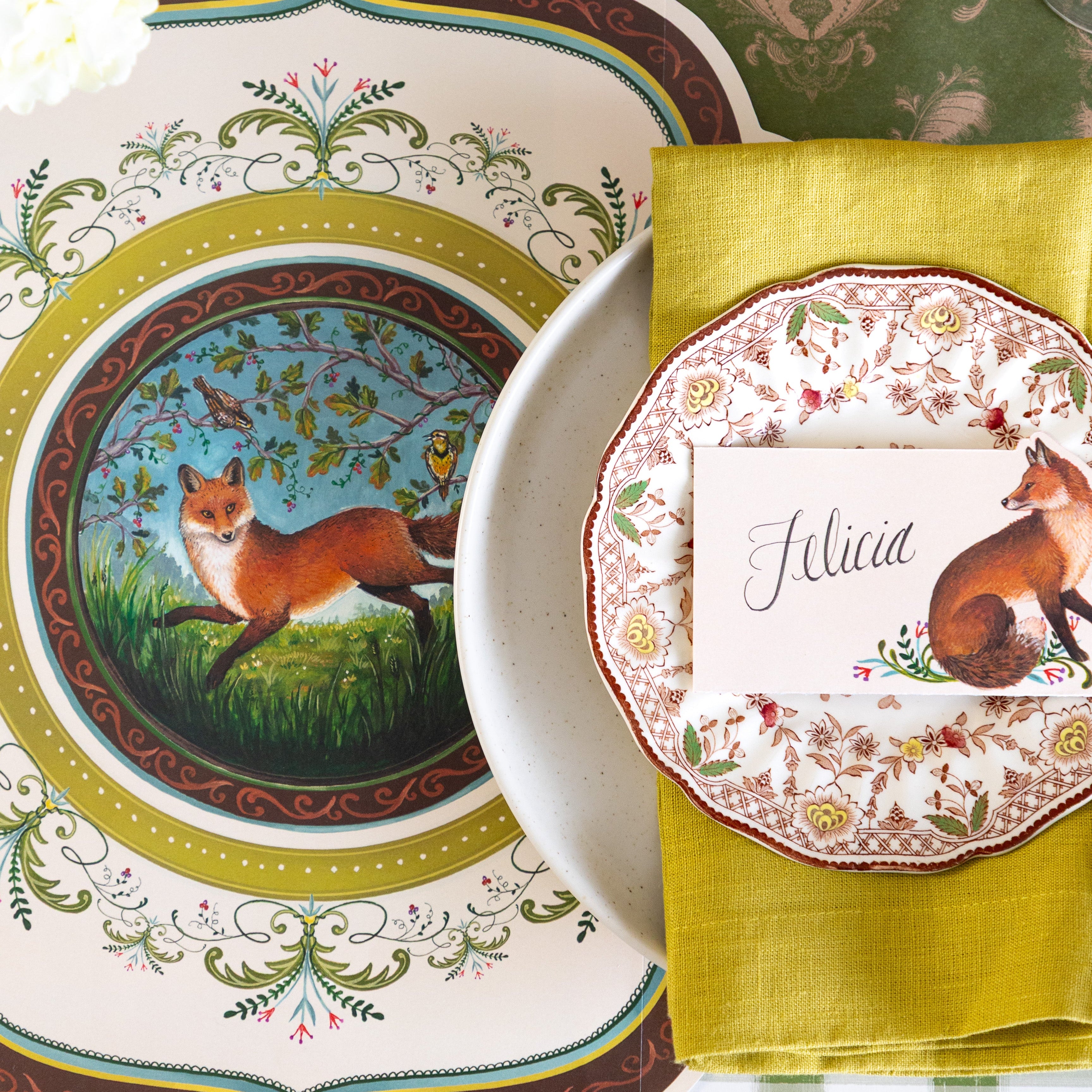 A decorative table setting featuring the Die-cut Fanciful Fox China Placemat, vintage dinnerware with the Fanciful Fox Place Card on top and Moss Fable Toile Runner underneath.