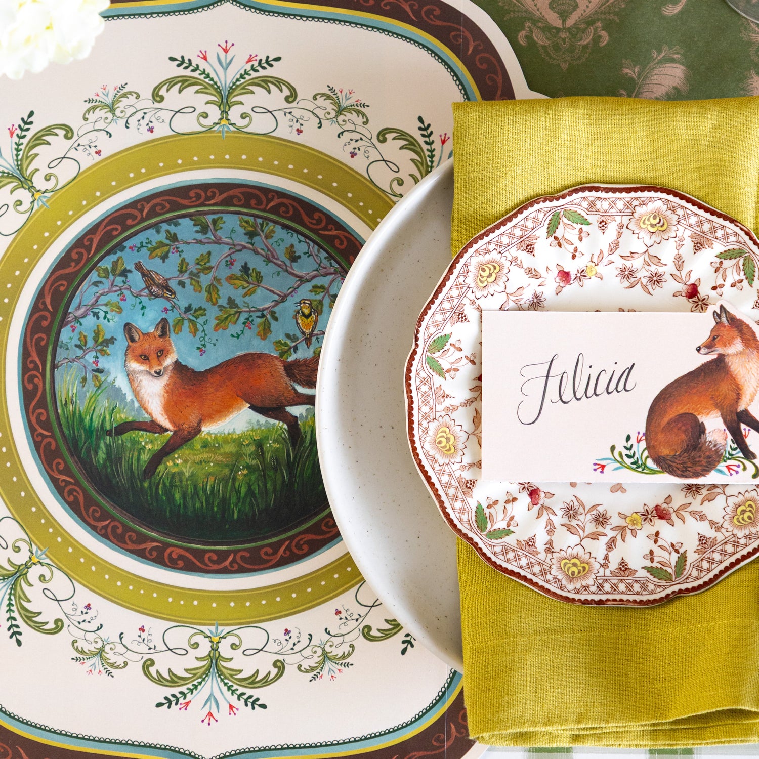 A decorative table setting featuring the Die-cut Fanciful Fox China Placemat, vintage dinnerware with the Fanciful Fox Place Card on top and Moss Fable Toile Runner underneath.