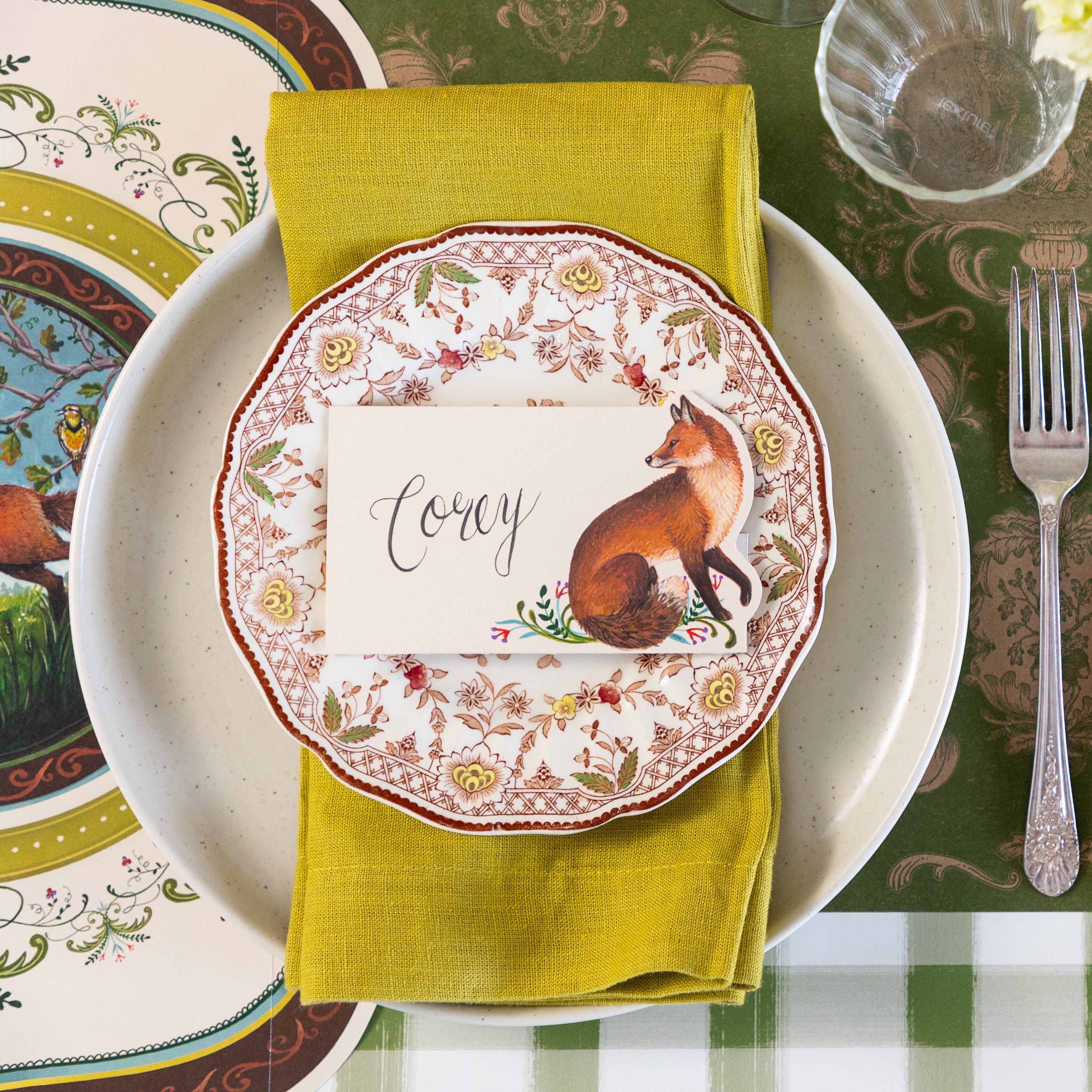 An elegant place setting featuring the Die-cut Fanciful Fox Placemat under a green napkin in between the Pacifica Salt Dinner plate and vintage floral salad plate with the Fanciful Fox Place Card reading "Corey" on top.