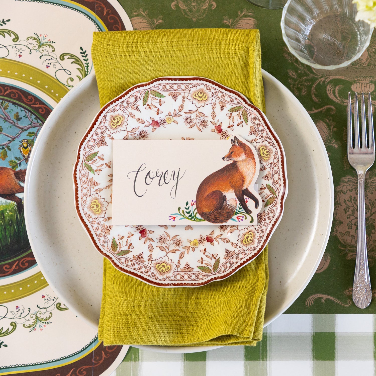 An elegant place setting featuring the Die-cut Fanciful Fox Placemat under a green napkin in between the Pacifica Salt Dinner plate and vintage floral salad plate with the Fanciful Fox Place Card reading &quot;Corey&quot; on top.