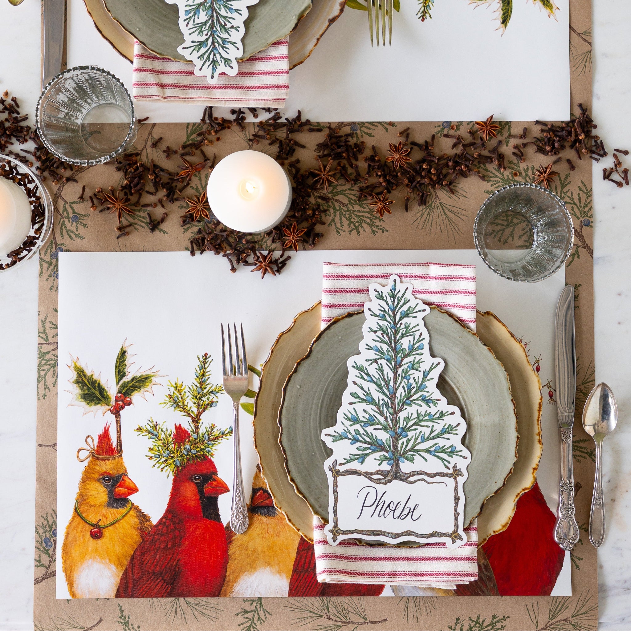 A holiday table setting features the Cardinal Party Placemat, woodland dinner plates, a red-and-white striped napkin, and a Cedar Tree Table Accent on the Juniper Sprigs Runner. Candles and star anise complete the festive holiday table decor.