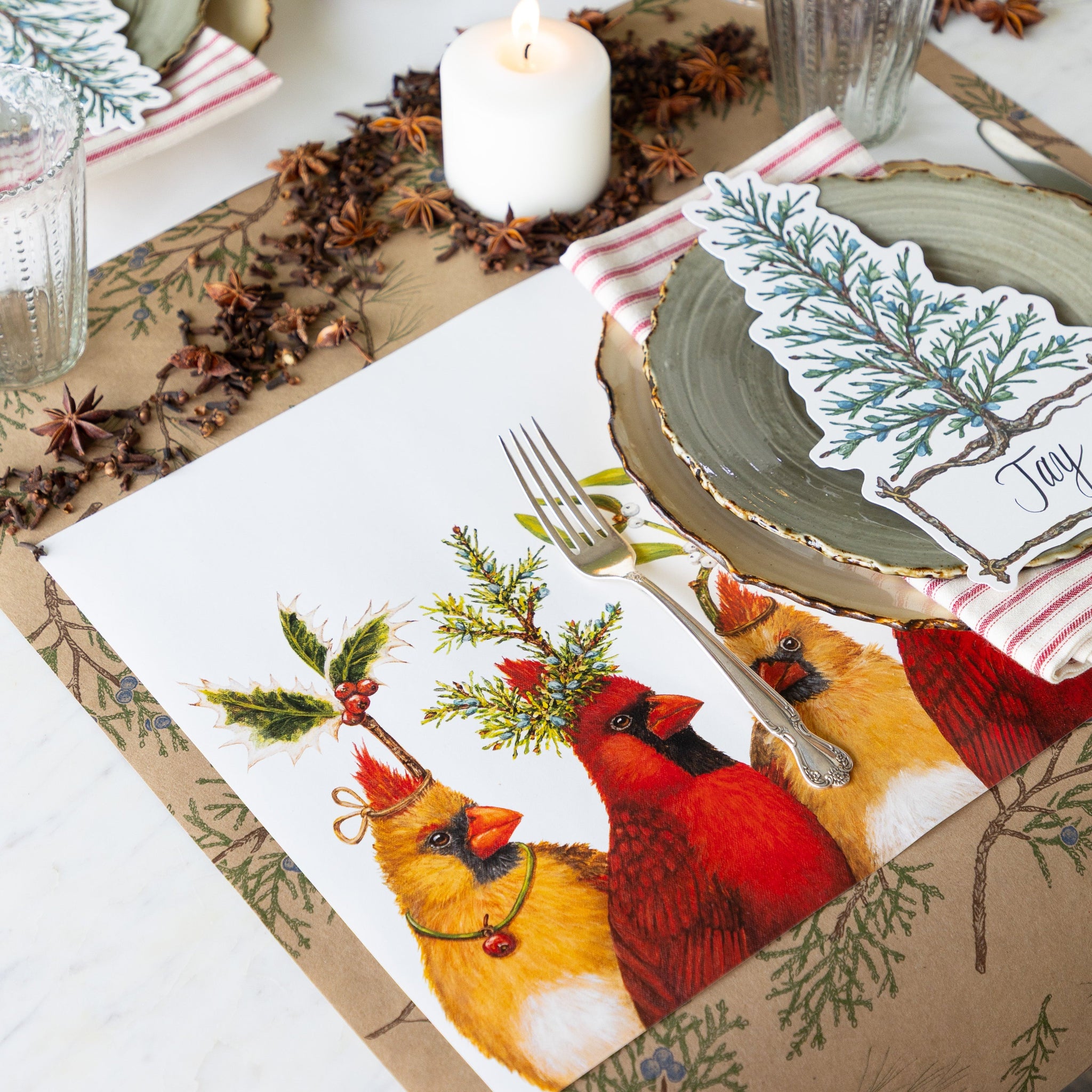A holiday table setting features the Cardinal Party Placemat, woodland dinner plates, a red-and-white striped napkin, and a Cedar Tree Table Accent on the Juniper Sprigs Runner. Candles and star anise complete the festive holiday table decor.