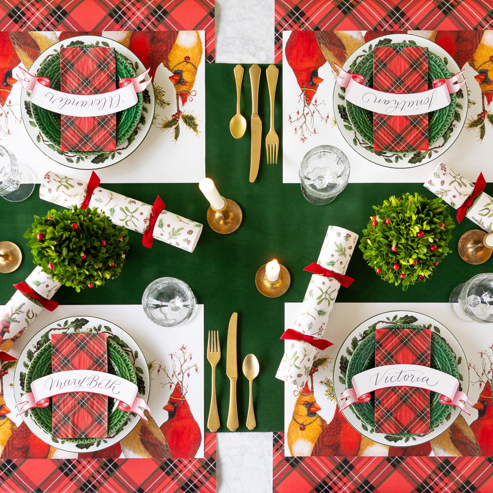 A festive holiday table setting features Cardinal Party Placemats, Red Plaid Napkins and runners, gold cutlery, topiary centerpieces, Cardinal Party Festive Crackers, candles, and Red Classic Banner Table Accents atop the Green Flocked Runner.