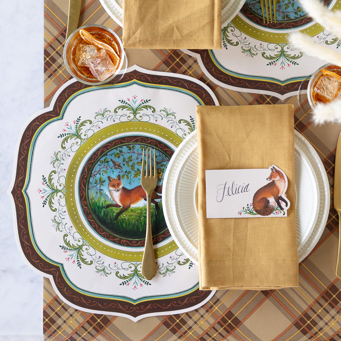 A decorative table setting featuring the Die-cut Fanciful Fox China Placemat, white dinnerware, tan napkin with the Fanciful Fox Place Card reading "Felicia", on top and Autumn Plaid runner underneath.