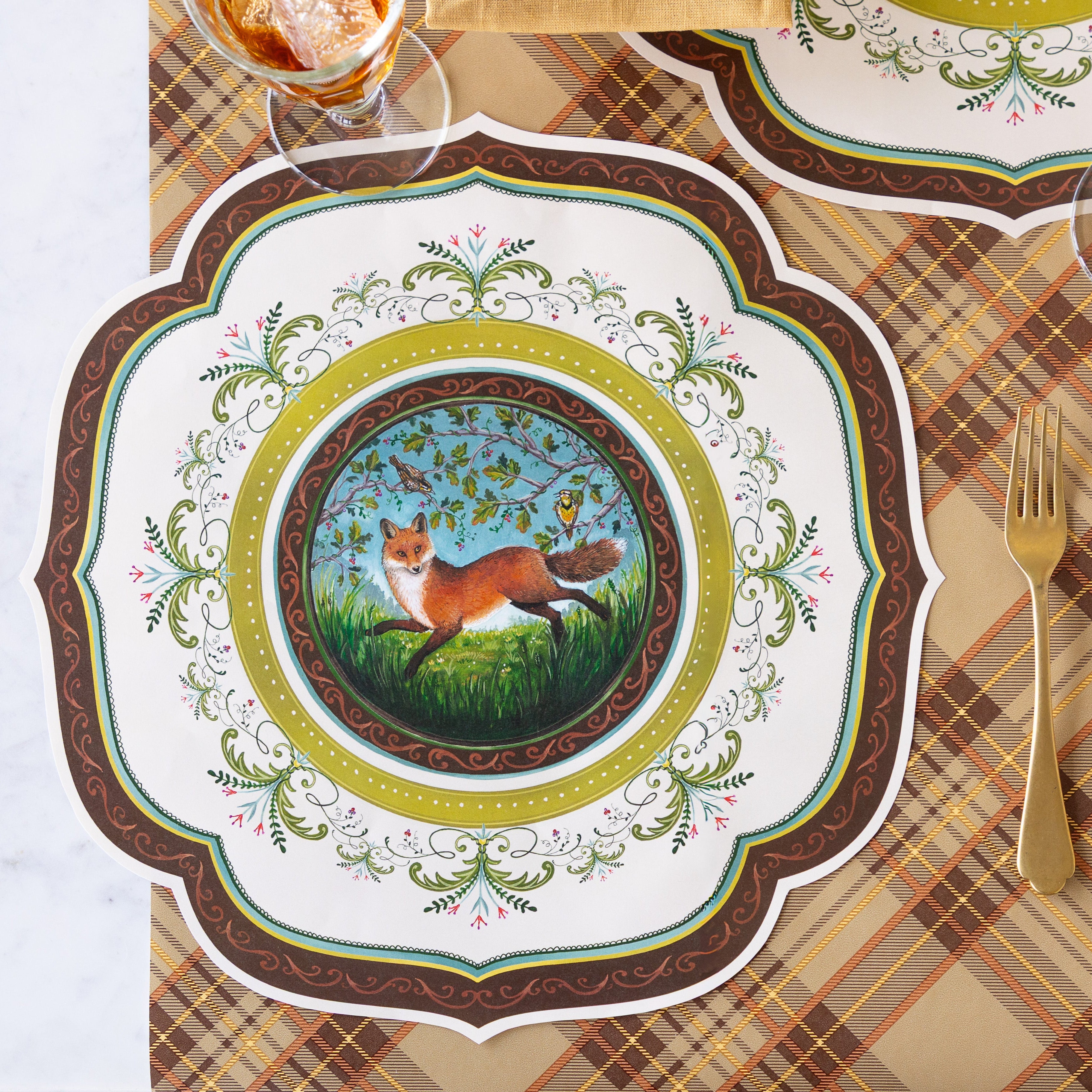 The Die-cut Fanciful Fox China Placemat on a table with a gold fork beside it, a glass of iced tea, and the Autumn Plaid runner underneath.