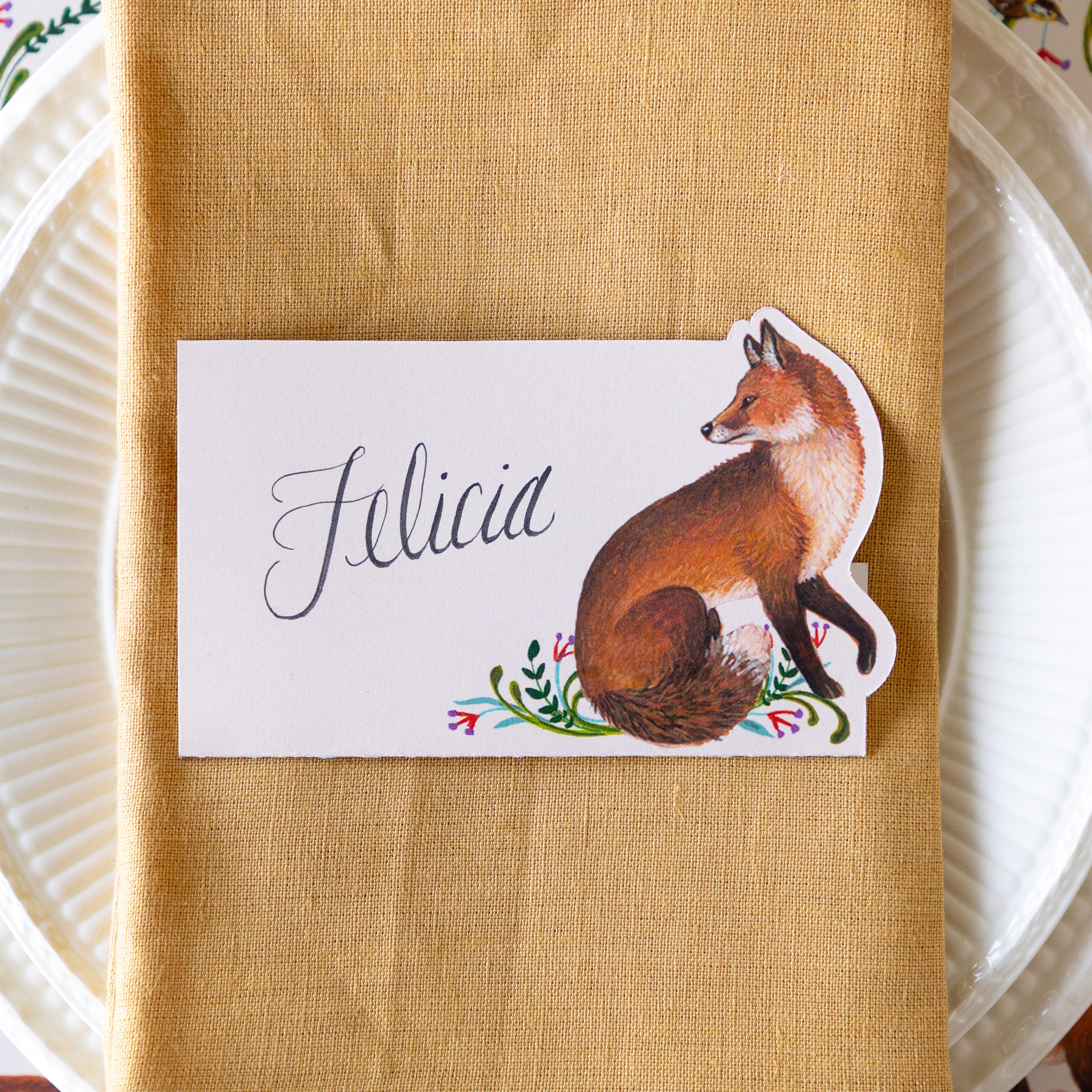 The Fanciful Fox Place Card reading "Felicia" on top of a tan plate and white dinner plates.