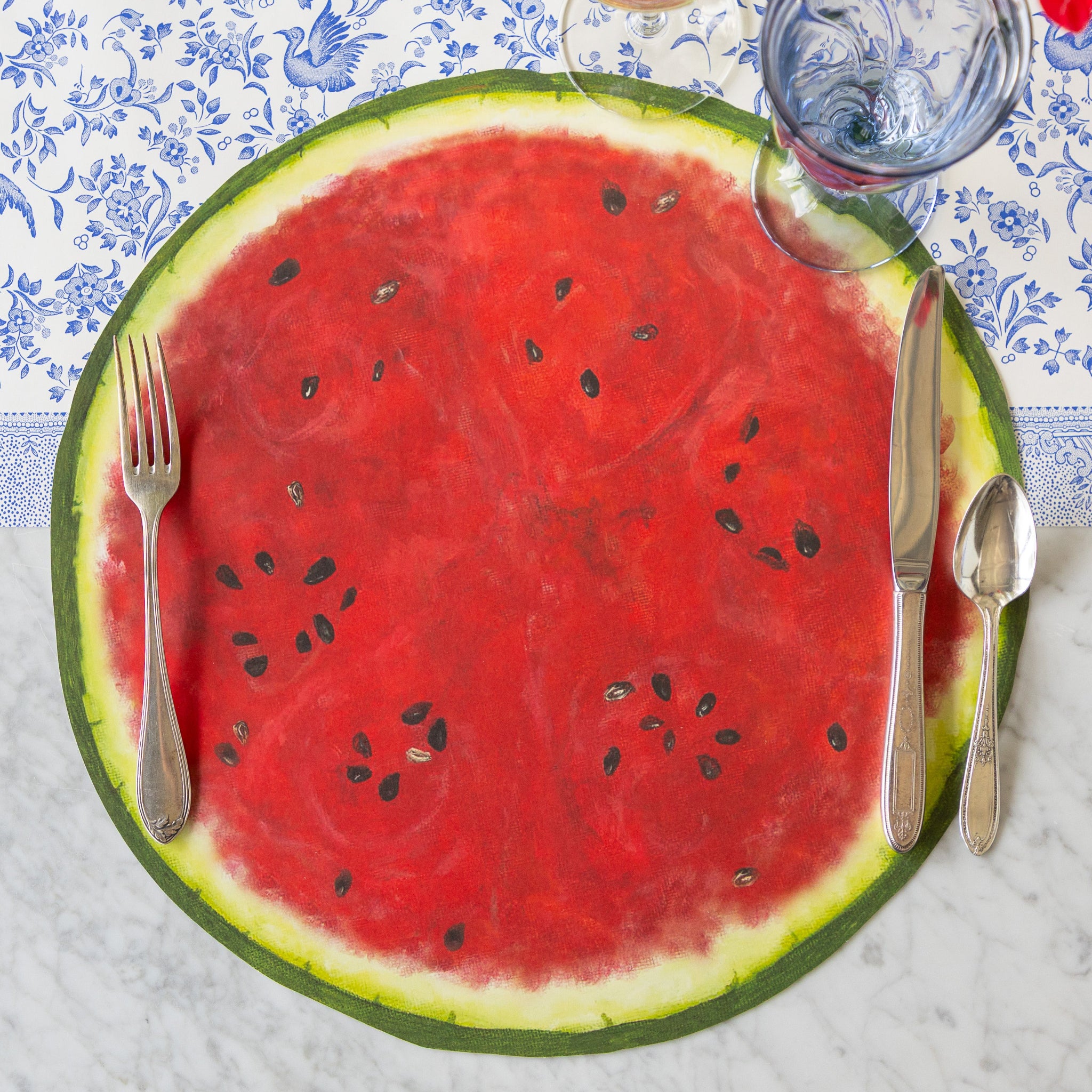 The Die-cut Watermelon Placemat with vintage silverware and a blue glass atop on the Blue Regal Peacock Runner.