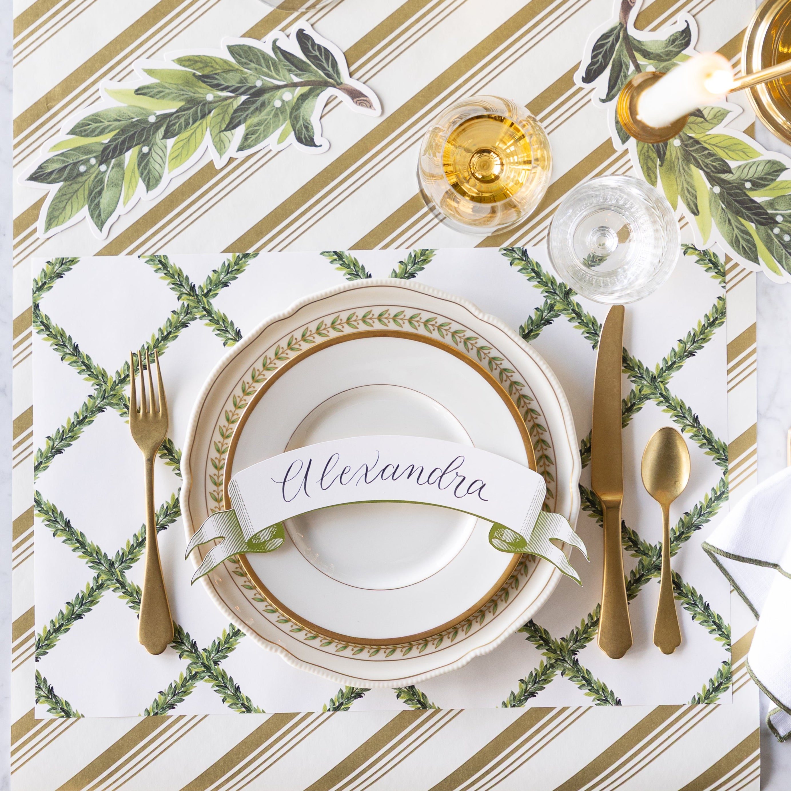 A formal place setting with gold cutlery, a wine glass, vintage plates with the Classic Moss Banner Table Accent reading &quot;Alexandra&quot; atop, Laurel Table Accents scattered on the table, and Laurel Wreath Placemats on the Gold Peppermint Stripe Runner.