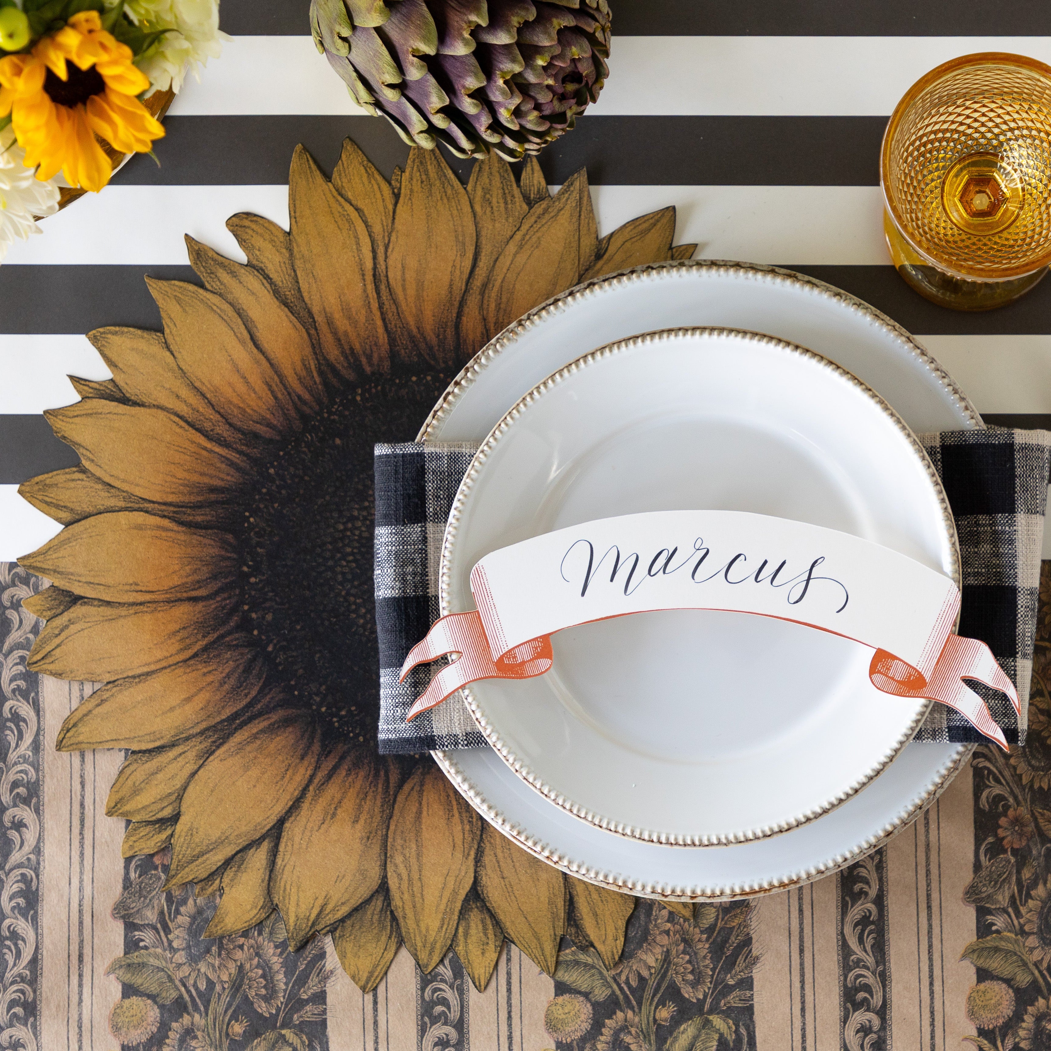 A place setting with fresh flowers and an artichoke, featuring the Die-cut Vintage Sunflower Placemat, a black checkered napkin, two white plates with the Classic Orange Banner Table Accent reading "Marcus" atop, Vintage Black Sunflower Runner rolled out vertically and Black Classic Stripe Runner horizontally underneath.