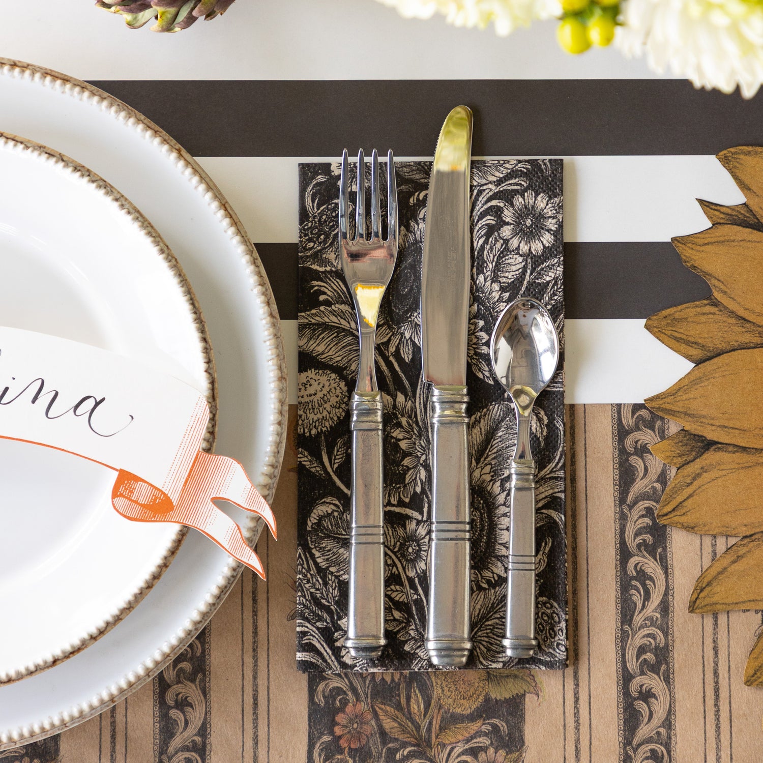 A place setting with the Vintage Sunflower Guest Napkin under silverware next to it,  on top of the Vintage Black Sunflower Runner and Black Classic Stripe Runner.