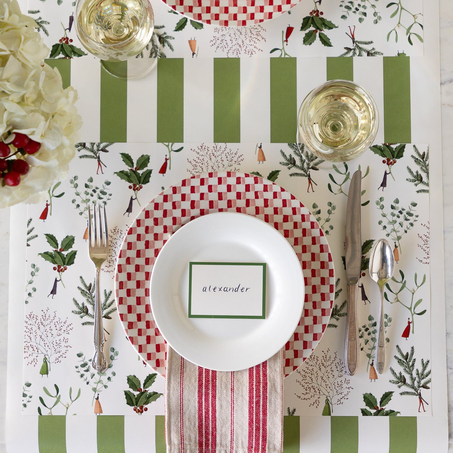 An elegant place setting featuring the Happy Christmas Placemat with a red checkered and white plate with the Dark Green frame Place Card reading &quot;alexander&quot; atop, vintage silverware, a glass of wine, and green stripe runner underneath.