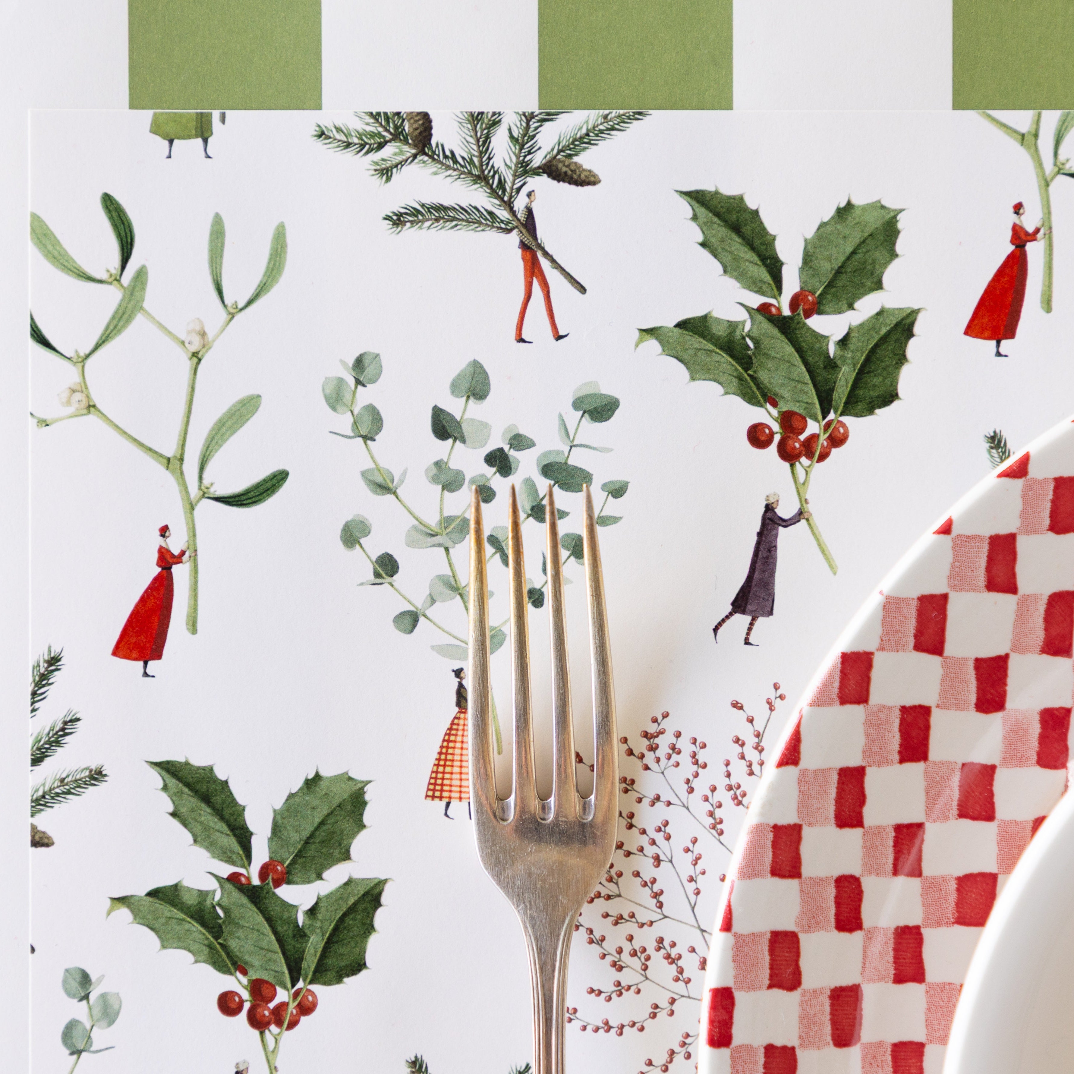 A silver fork rests on the Happy Christmas Placemat with illustrated tiny figures holding large greenery, holly and eucalyptus, next to a red-and-white checkered plate and striped napkin atop.