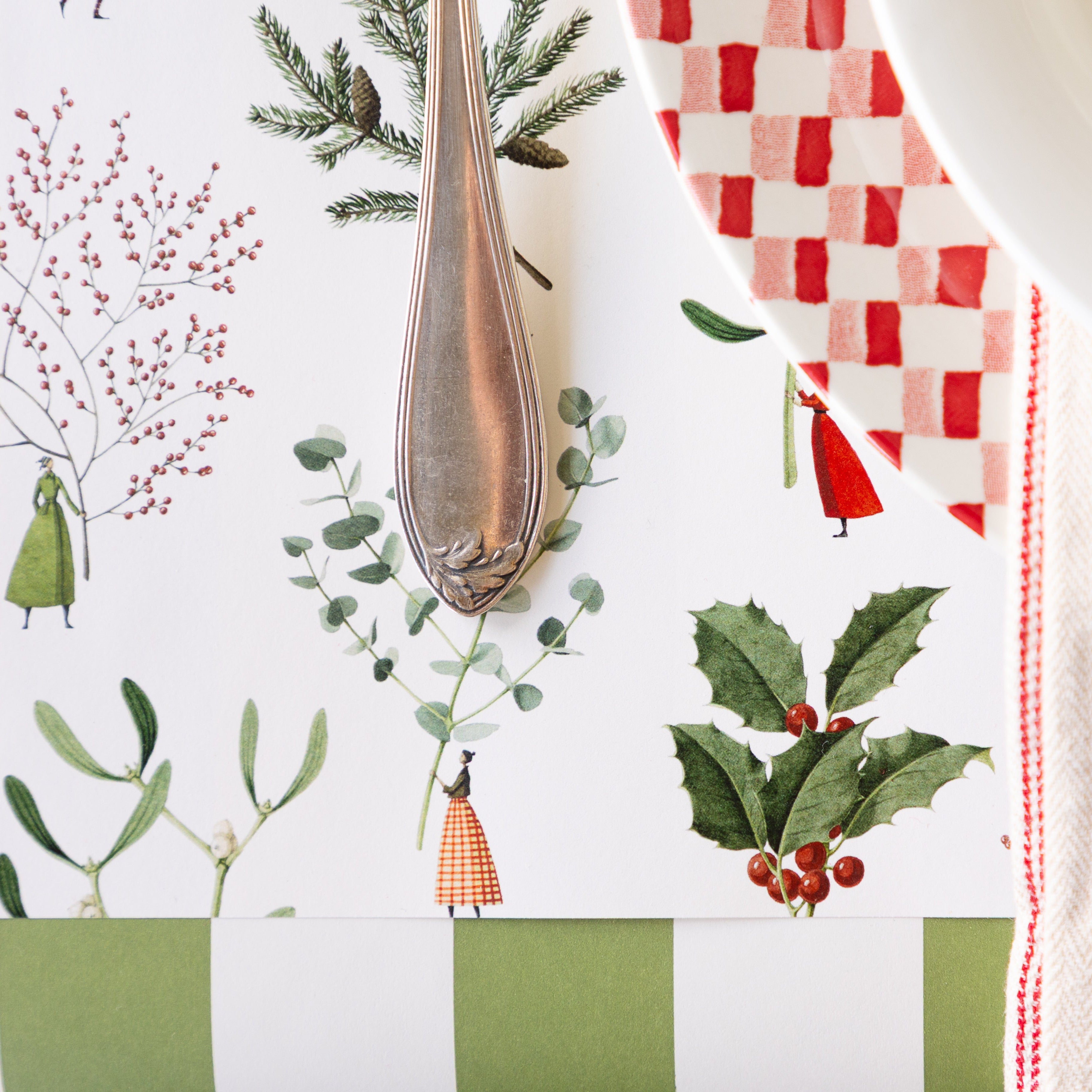 A silver fork rests on the Happy Christmas Placemat with illustrated tiny figures holding large greenery, eucalyptus and holly, next to a red-and-white checkered plate and striped napkin atop.