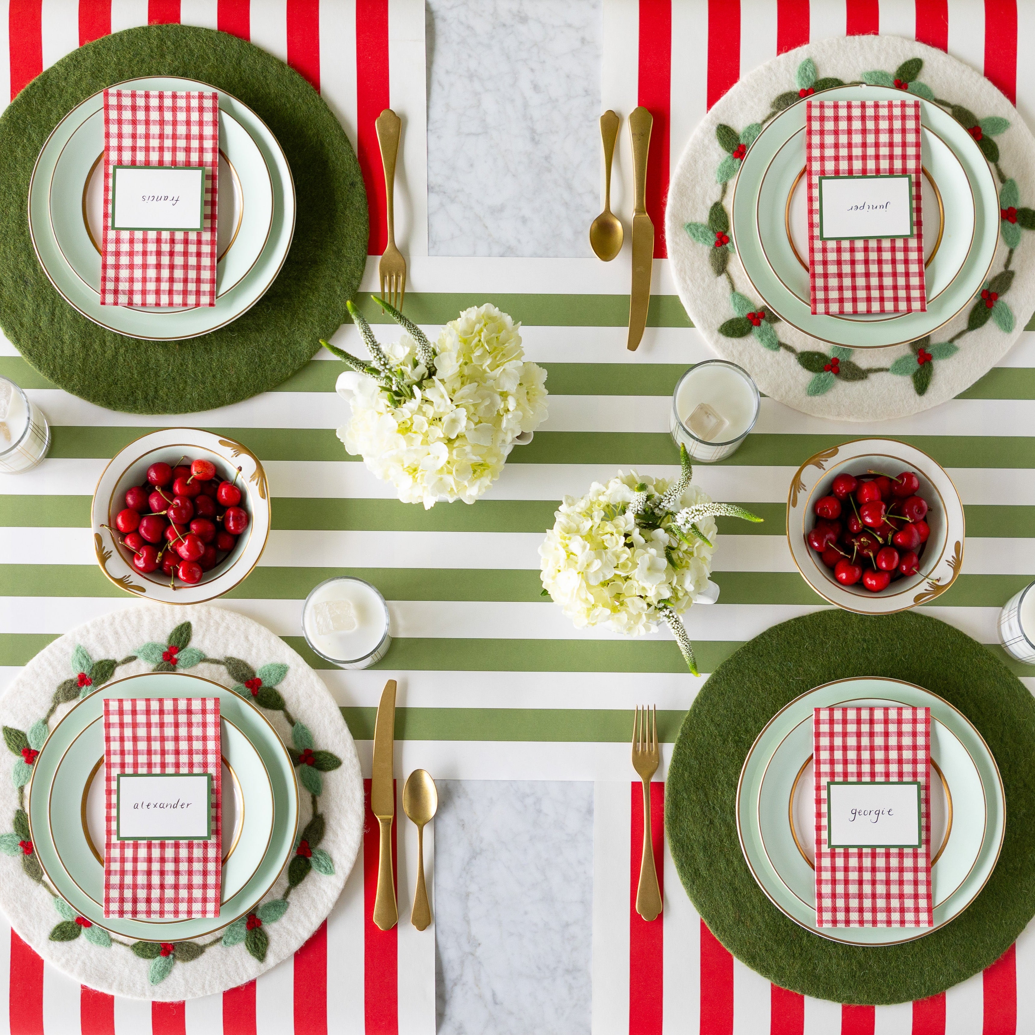 A table for four is set with flowers, Moss Green and Berry Wreath Felt Placemats, seafoam plates with Red Painted Check Guest Napkins and Dark Green Frame Place Cards atop, glasses of milk. gold flatware, and bowls of cherries on the Red Classic and Moss Green Classic Stripe Runners.