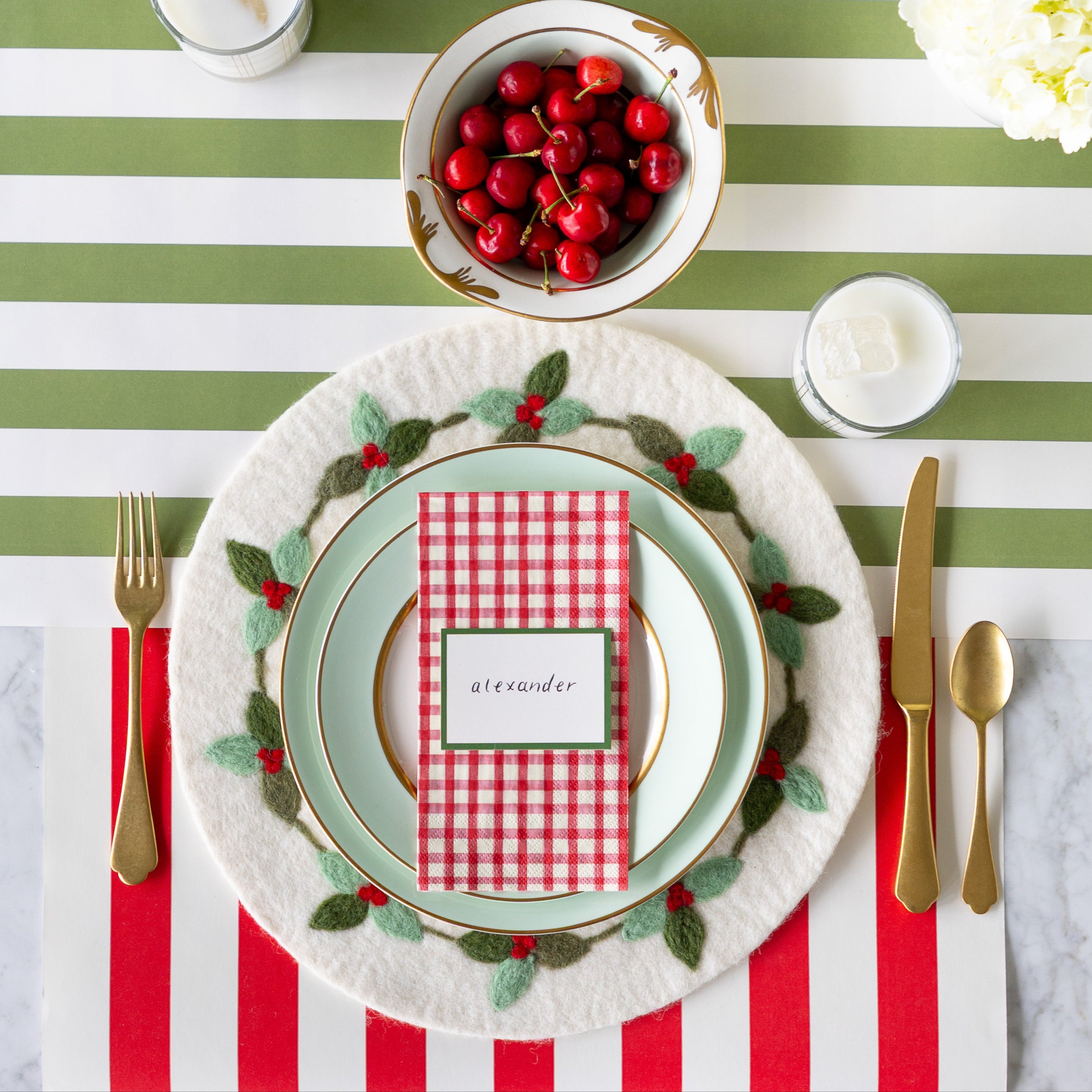 An elegant place setting featuring the Felt Mistletoe Berry Placemat, seafoam dinnerware with the Red Plaid Guest Napkin and Dark Green Frame Place Card atop, gold flatware, a glass of milk and bowl of cherries on the Red Classic Stripe and Moss Classic Stripe Runners.