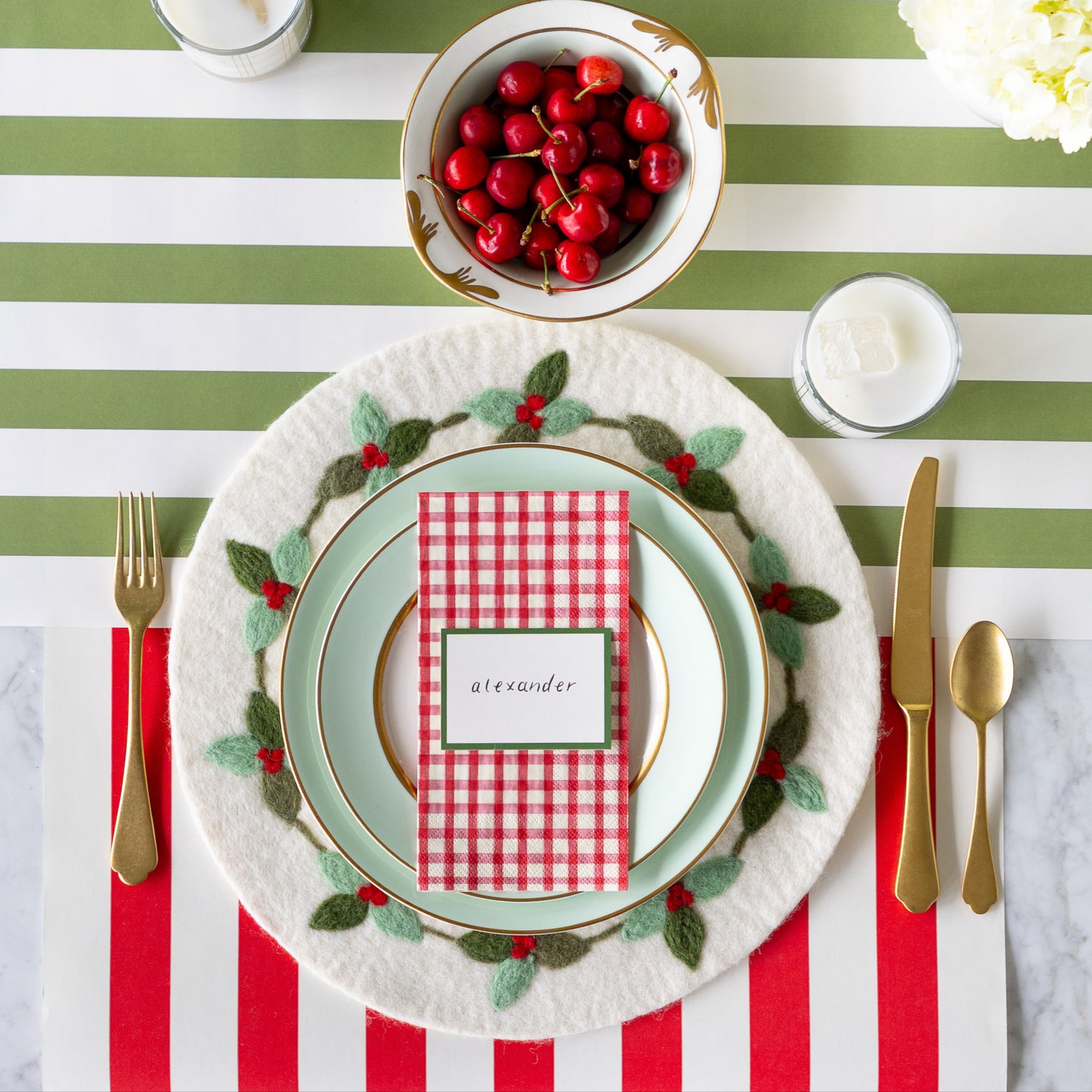 An elegant place setting featuring the Felt Mistletoe Berry Placemat, seafoam dinnerware with the Red Plaid Guest Napkin and Dark Green Frame Place Card atop, gold flatware, a glass of milk and bowl of cherries on the Red Classic Stripe and Moss Classic Stripe Runners.