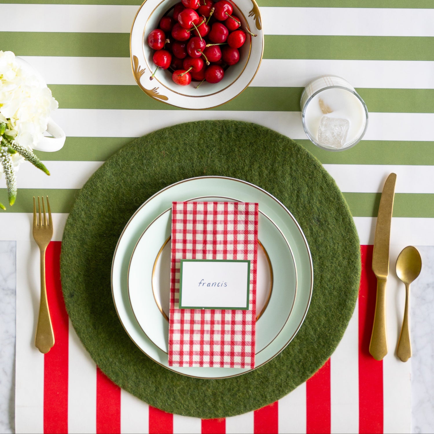 A place setting featuring the Forest Green Felt Placemat, seafoam plates with the Red Painted Check Napkin and Dark Green Frame Place Card atop, gold flatware, a glass of milk and bowl of cherries on the Red Classic and Moss Classic Stripe Runners.