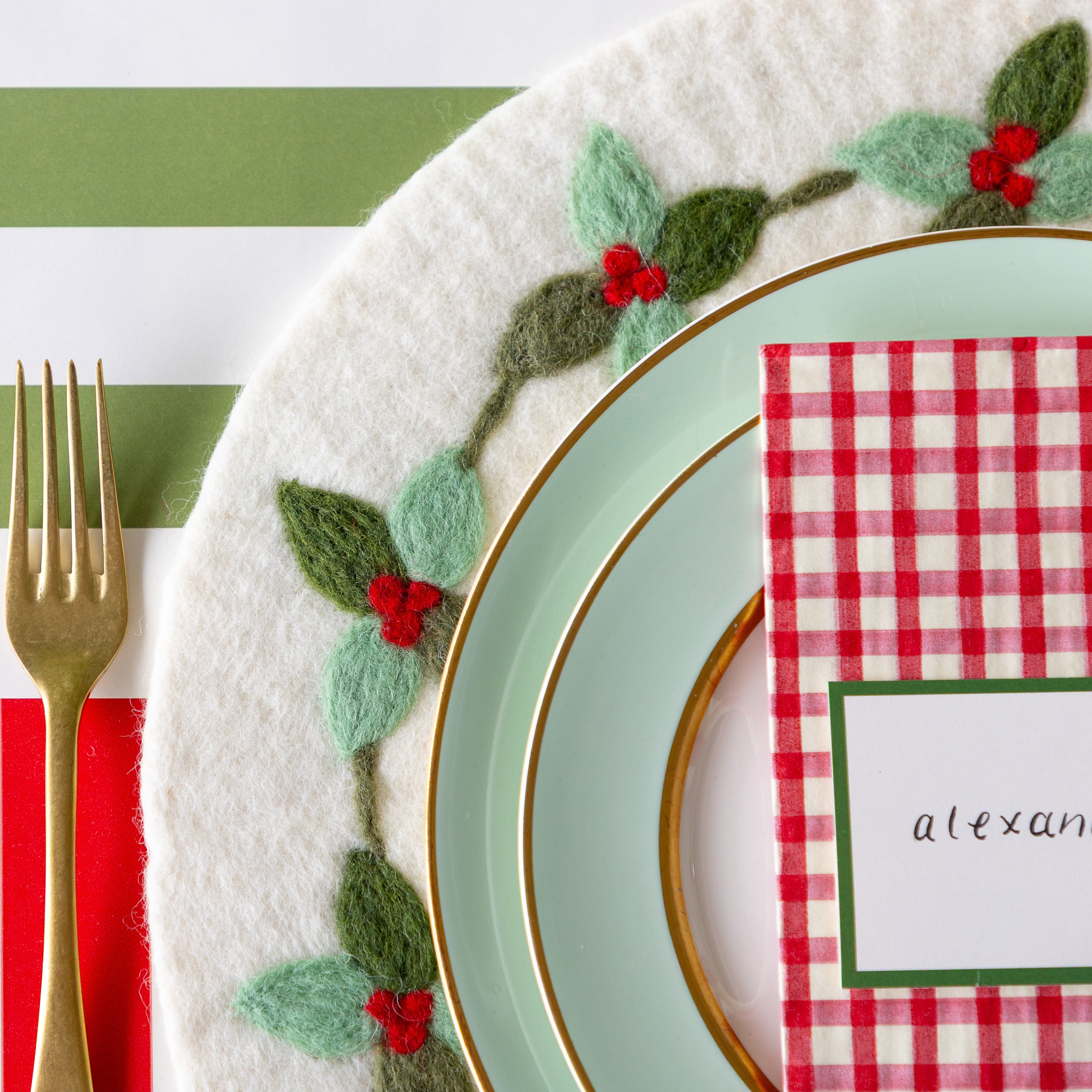 An elegant place setting featuring the Felt Mistletoe Berry Placemat, seafoam dinnerware with the Red Plaid Guest Napkin and Dark Green Frame Place Card atop, and gold flatware on the Red Classic Stripe and Moss Classic Stripe Runners.