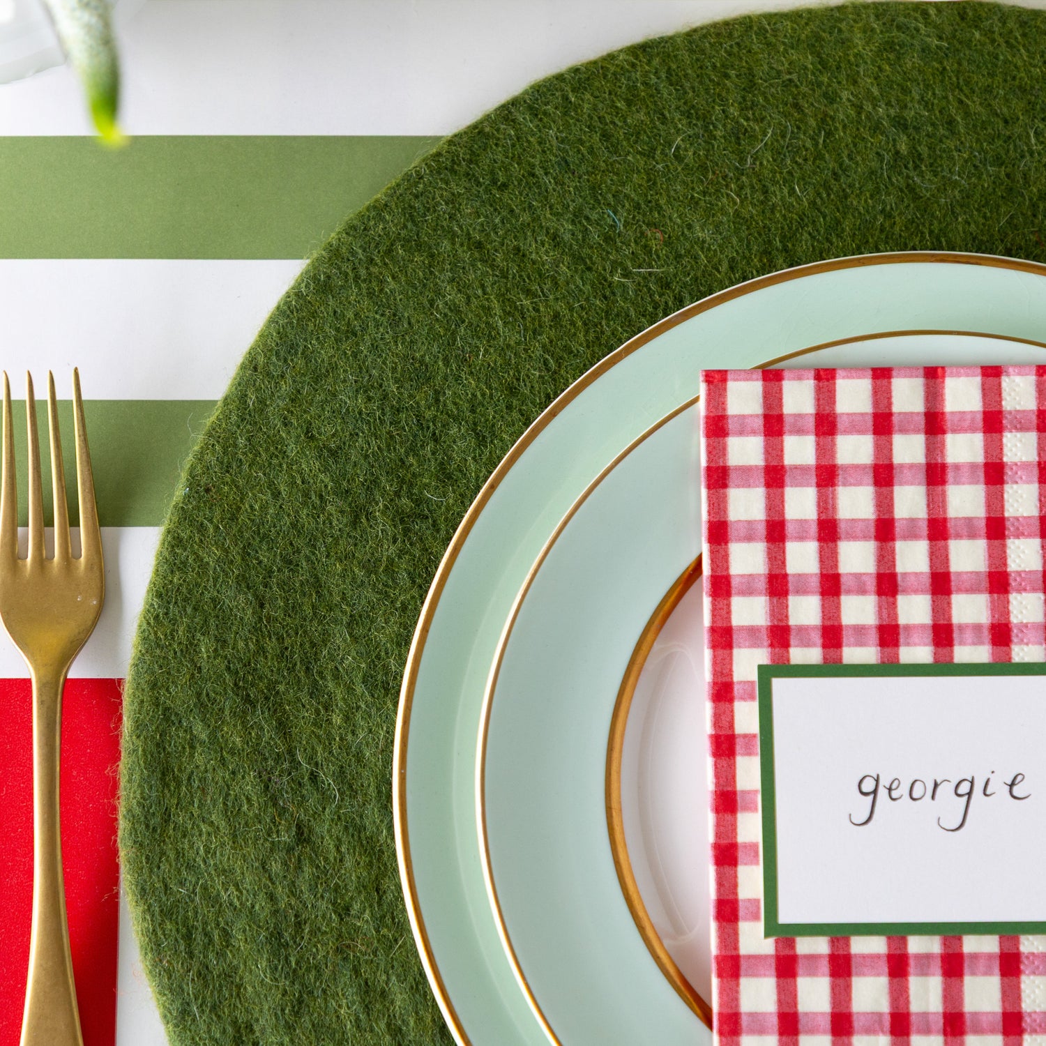 A place setting featuring the Forest Green Felt Placemat, seafoam plates with the Red Painted Check Napkin and Dark Green Frame Place Card atop and gold flatware on the Red Classic and Moss Classic Stripe Runners.